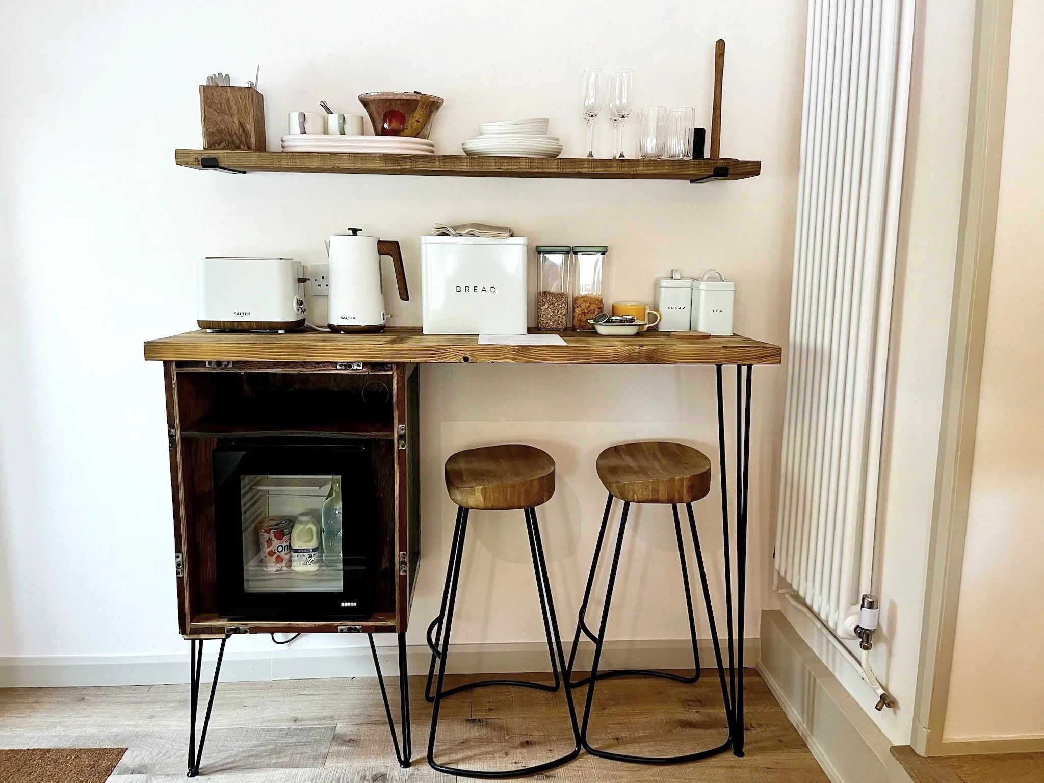 Kitchen or dining area with wooden shelves holding plates, glasses, bowls, and cups, a small fridge on a wooden stand containing beverages, and two wooden stools in front of a wall with a radiator and a power outlet.