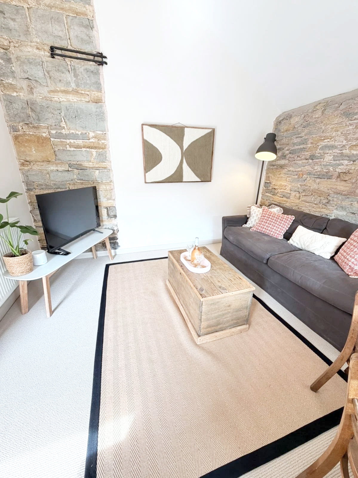Living room with a dark gray sofa, wooden coffee table, flat-screen TV on a white stand, potted plant, and a white wall decorated with a geometric art piece. Exposed brick wall sections on the left and right sides, beige rug with black border, and a 