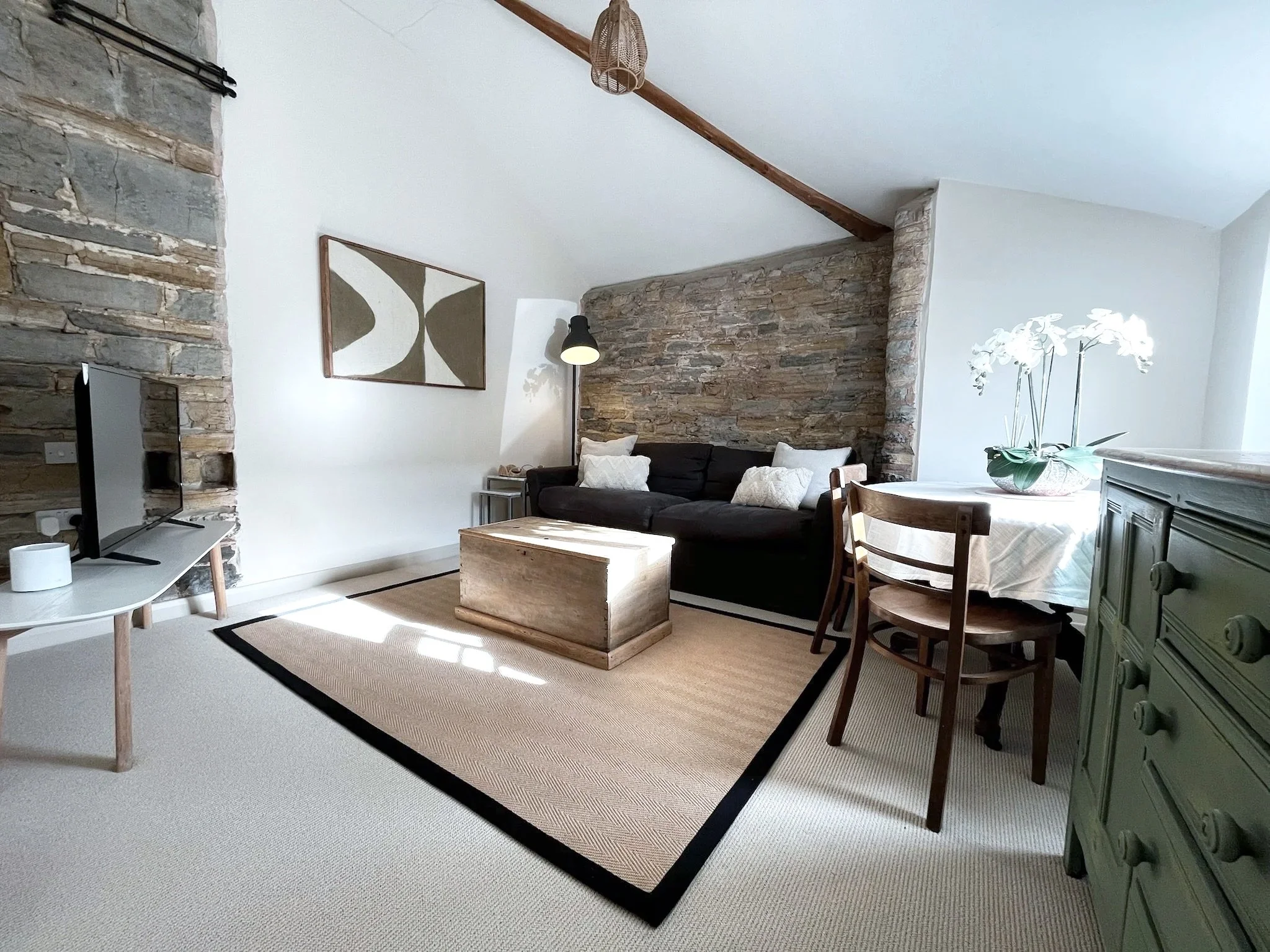 Living room with stone accent wall, a dark sofa with pillows, a wooden coffee table, a dining table with chairs, a green dresser, a potted orchid, and a wall art piece. The room has a mix of modern and rustic decor with natural light.