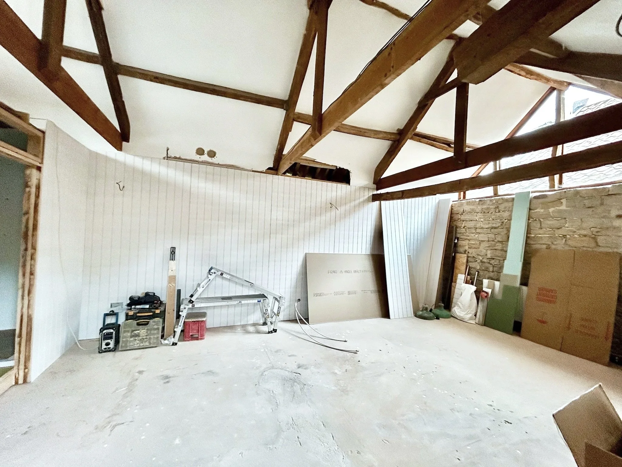 Room under renovation with exposed wooden beams on the ceiling, a partially finished wall with white paneling, construction tools, and materials scattered on the floor.