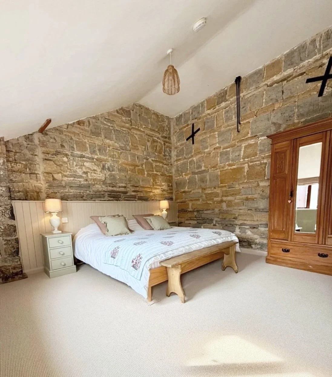 A bedroom with a stone wall, a bed with white bedding, bedside lamps, a wooden wardrobe with mirror, and beige carpet.