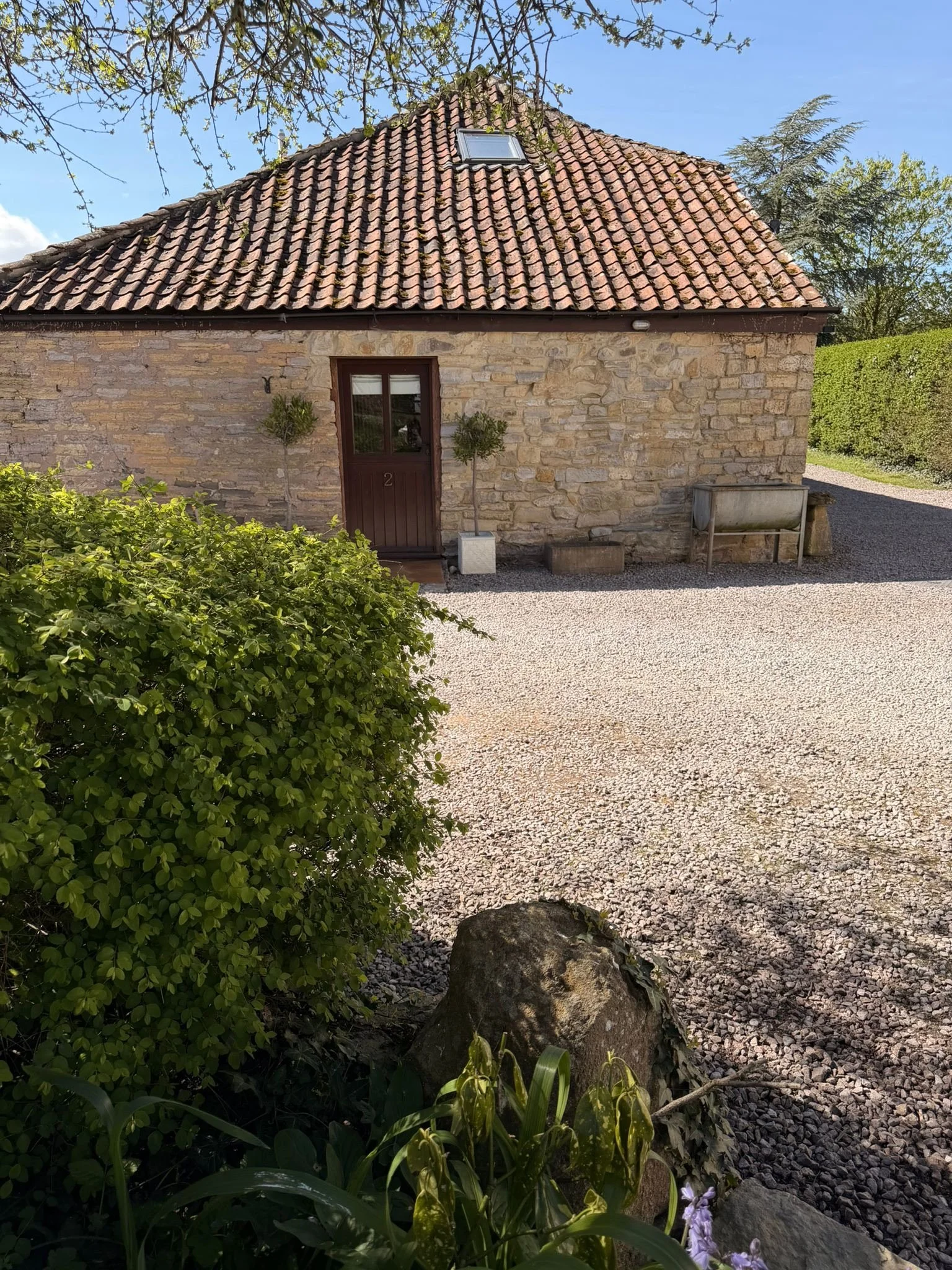 A stone house with a tiled roof, a small window, and a brown door, surrounded by greenery and gravel.