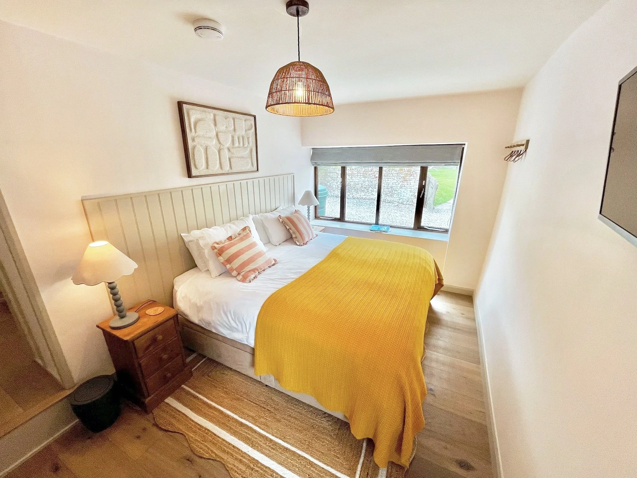 A bedroom with a bed covered with a yellow blanket, pink striped pillows, and white sheets. There is a wooden nightstand with a lamp, a large window with a gray shade, and artwork hanging above the bed. The room has wooden flooring and a striped rug.