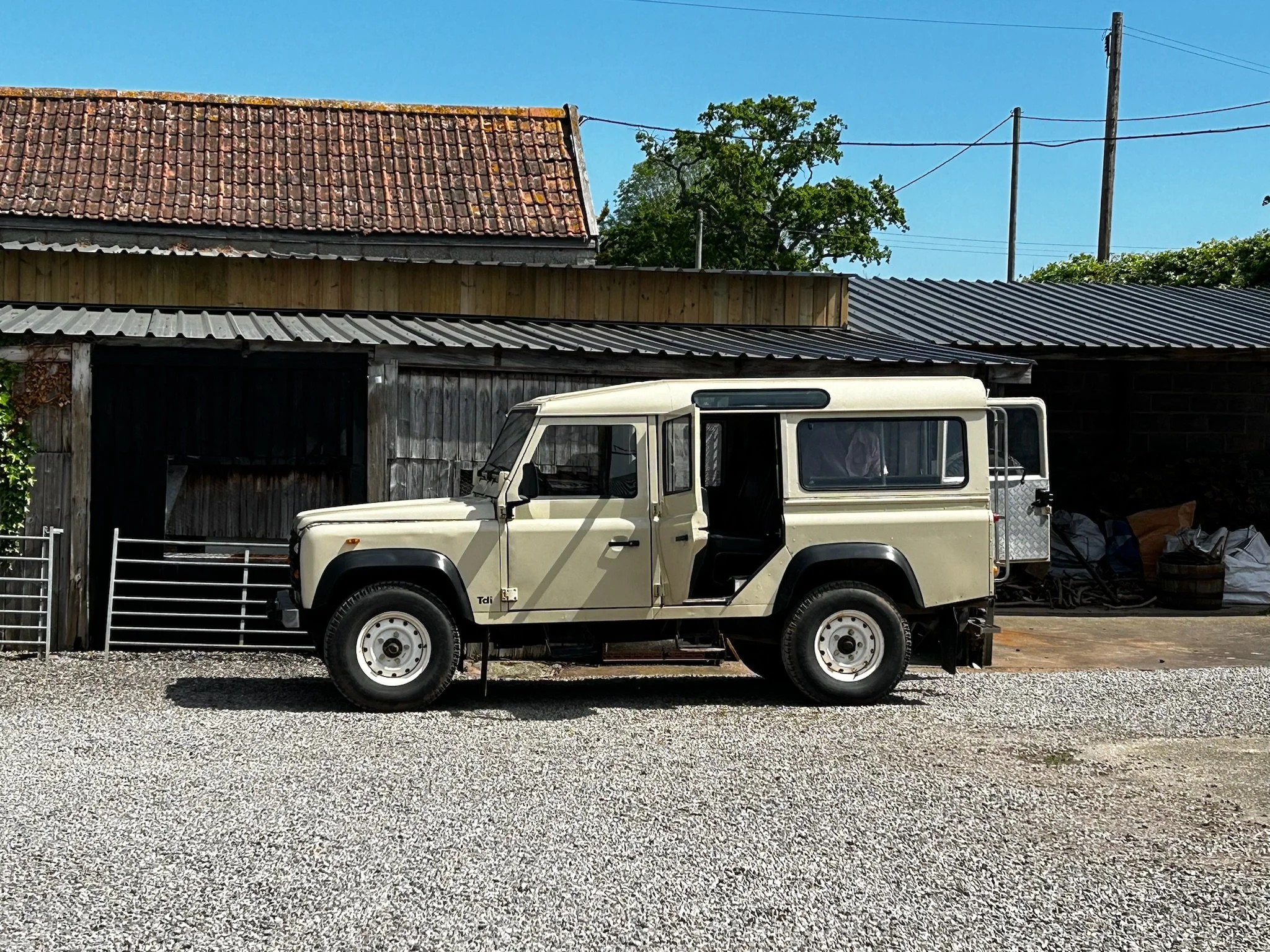 A vintage cream-colored off-road vehicle parked in front of a wooden building with a sloped metal roof. The building appears to be a barn or shed, with an area under construction or renovation. The background includes a clear blue sky, some trees, an