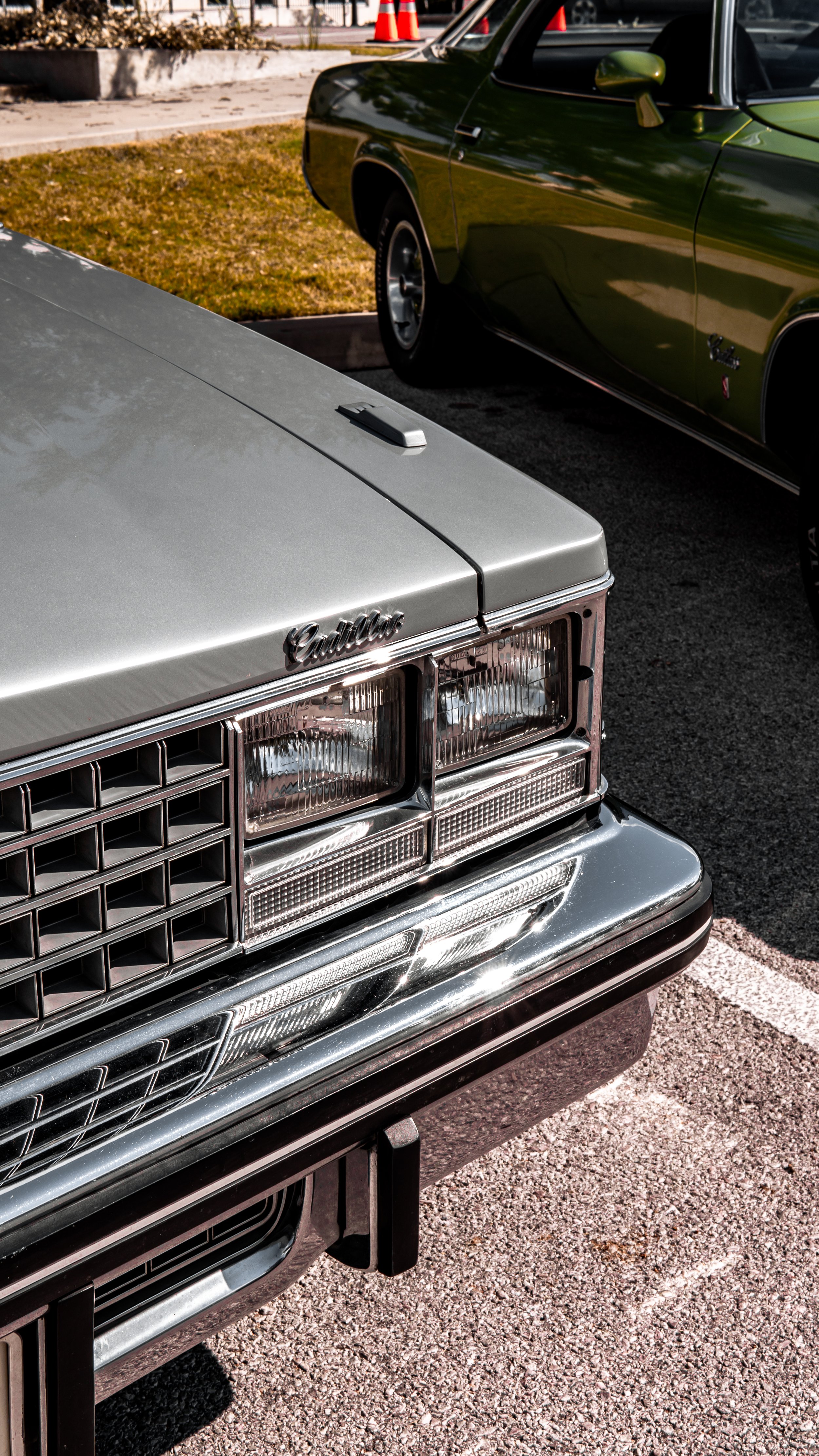 Primer plano de la parte frontal de un auto clásico plateado, con el nombre "Cadillac" en la parrilla y faros delanteros rectangular. Al fondo, un auto verde y conos de tráfico naranja.