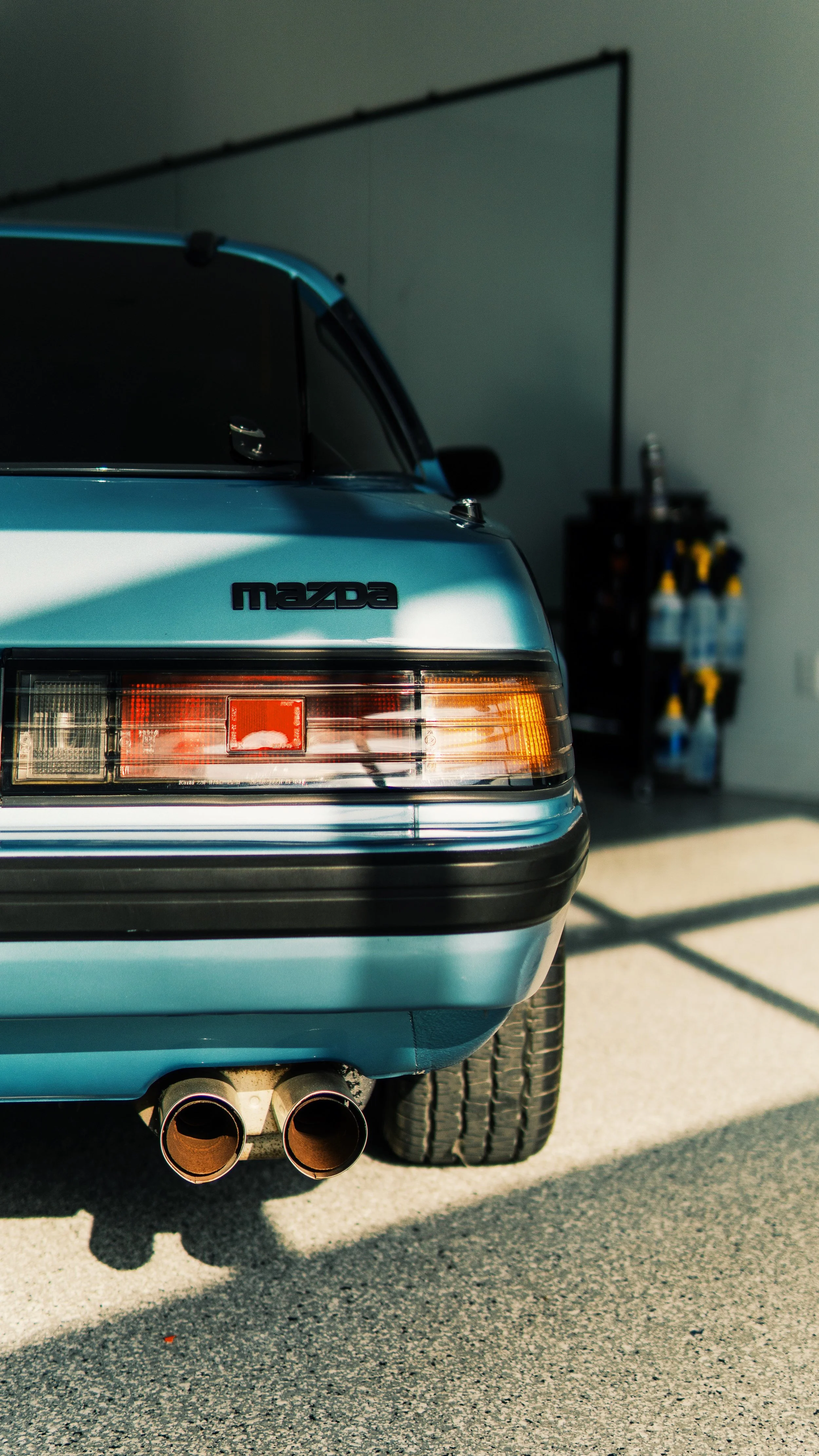 Parte trasera de un coche Mazda de color azul en un garaje, con doble tubo de escape y neumáticos.