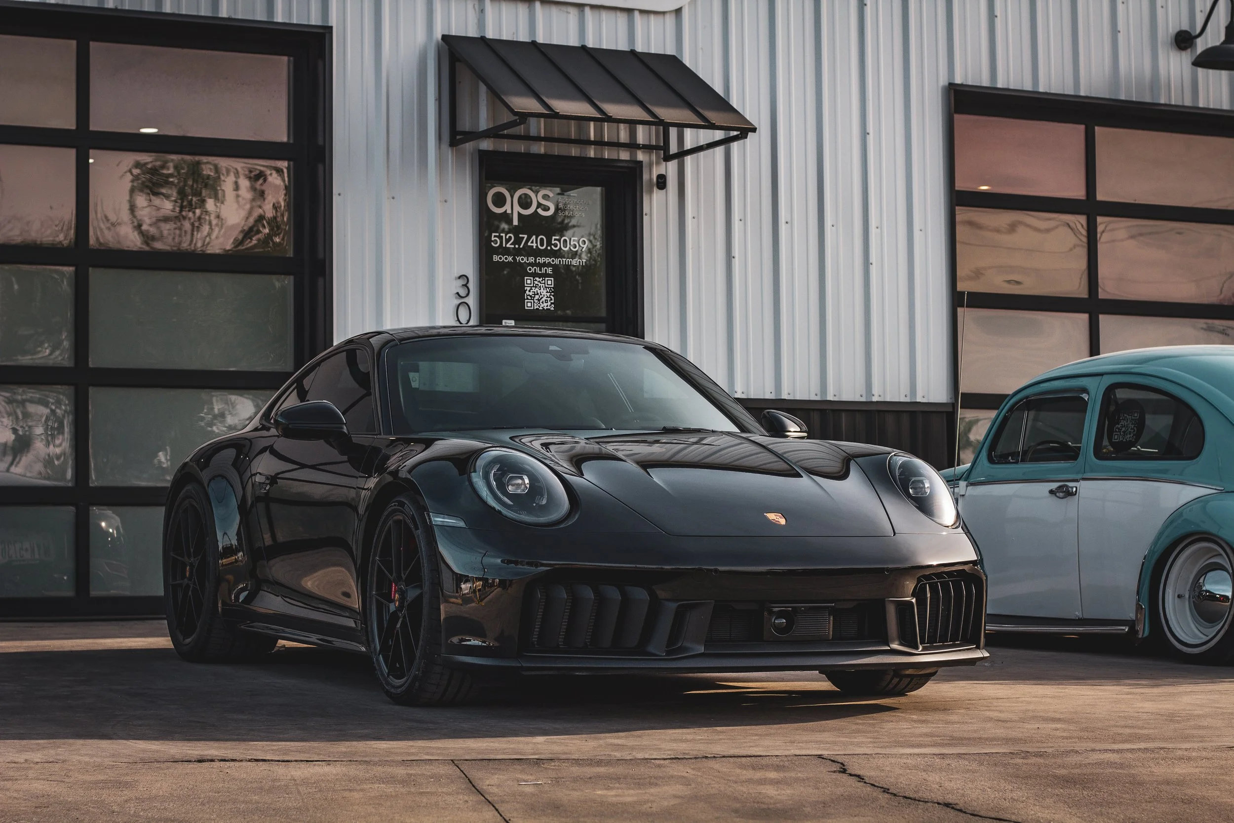Un auto deportivo Porsche negro estacionado frente a un edificio con puertas de vidrio, junto a un auto clásico blanco y verde.