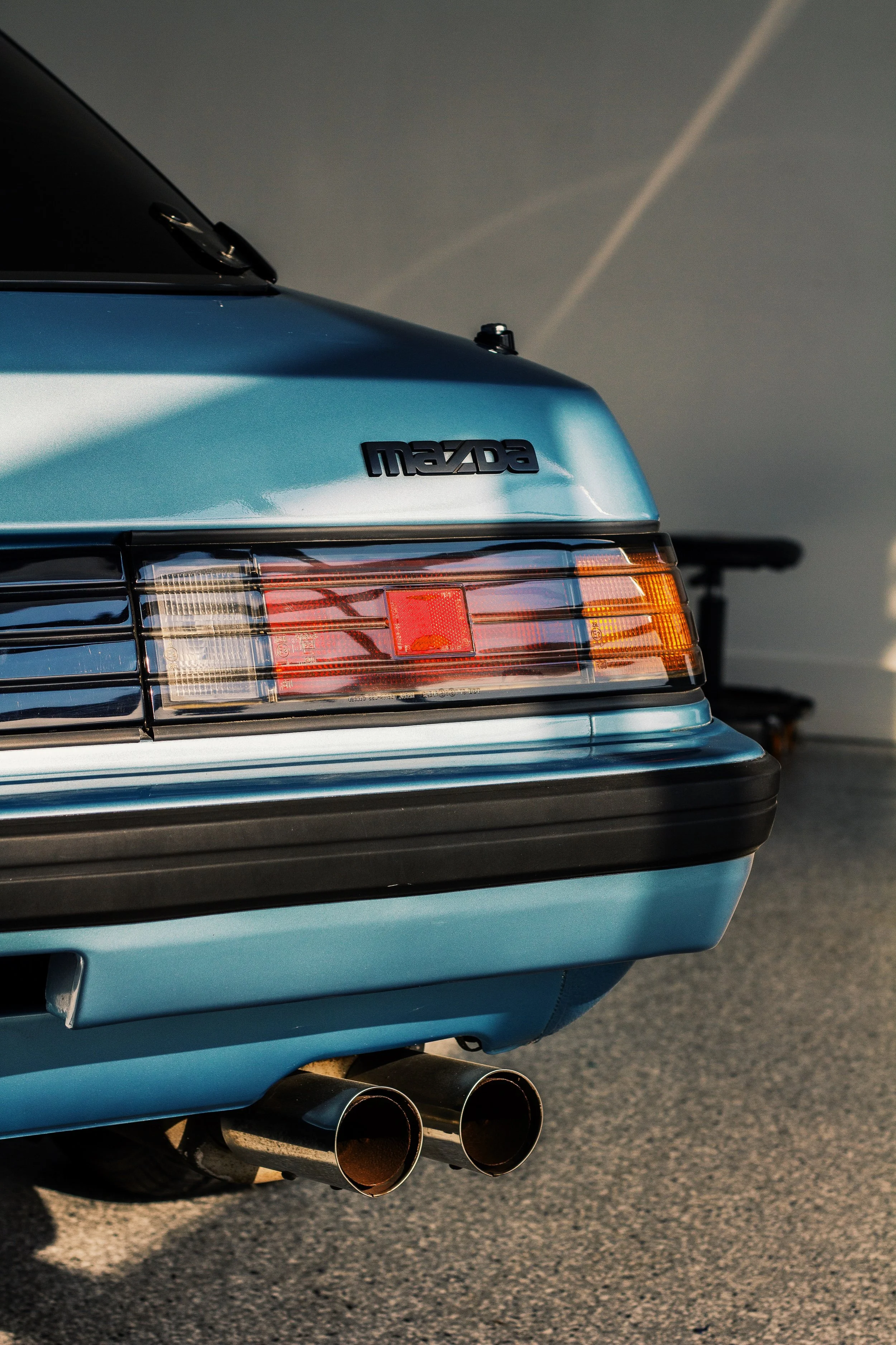 Parte trasera de un automóvil azul con emblema Mazda, luces traseras y doble tubo de escape en un pavimento.