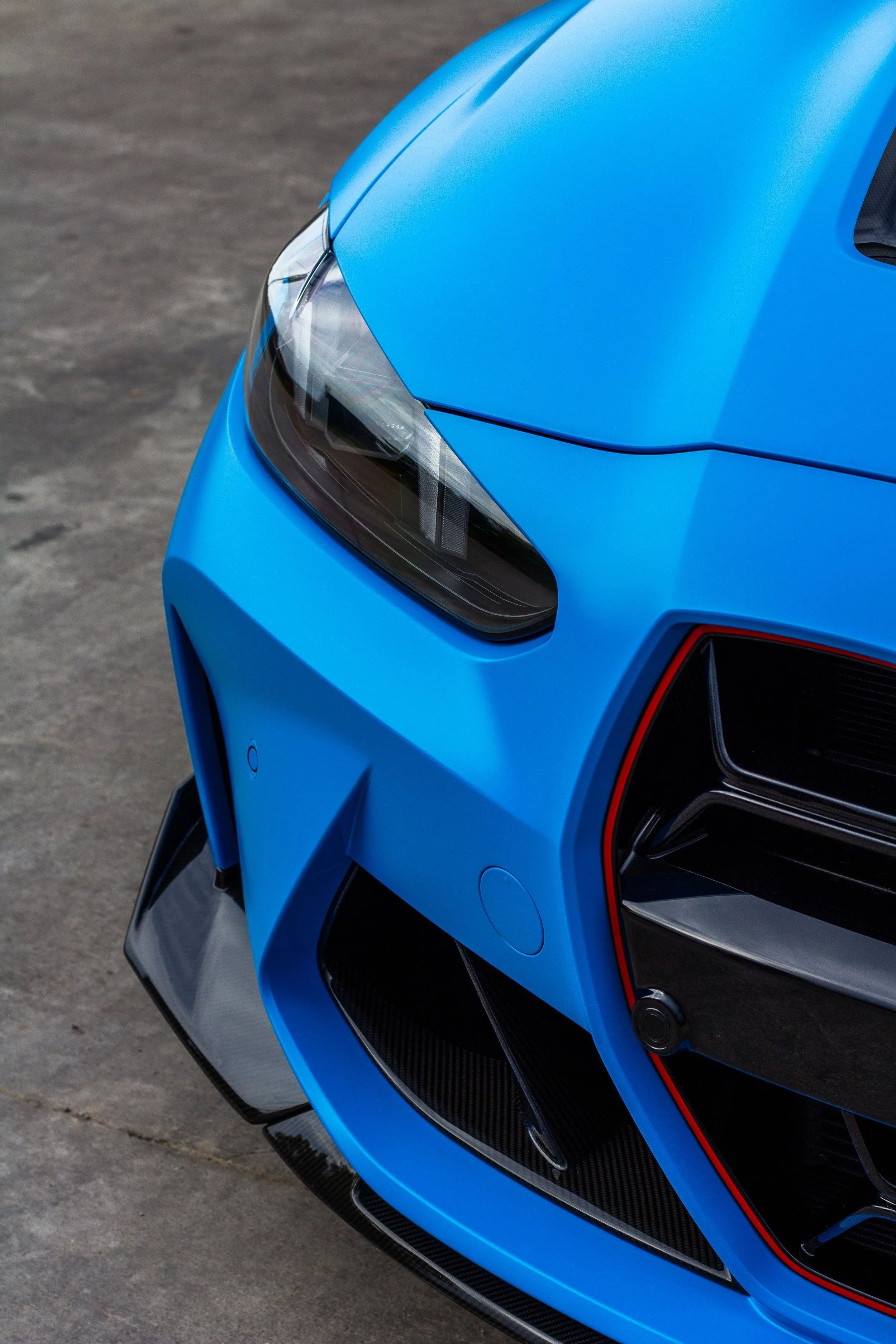 Primer plano del frente de un coche deportivo azul, mostrando la cabeza y el parachoques, con detalles de fibra de carbono y luces LED.