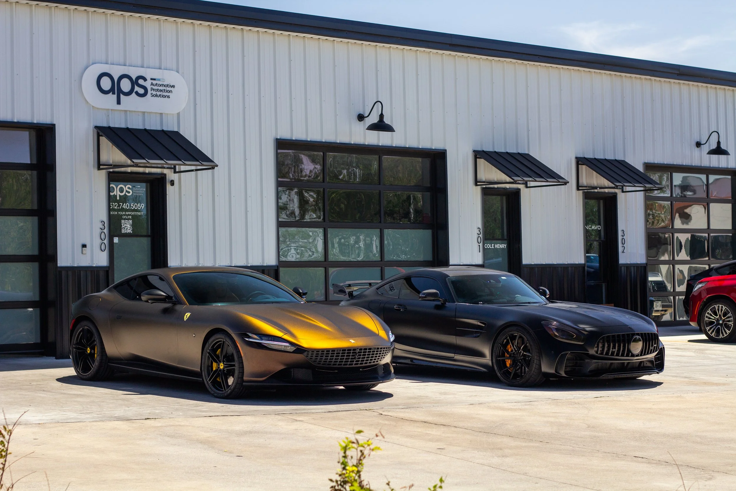Dos autos deportivos negros en un taller de reparación con un edificio de paredes blancas y ventanas grandes.