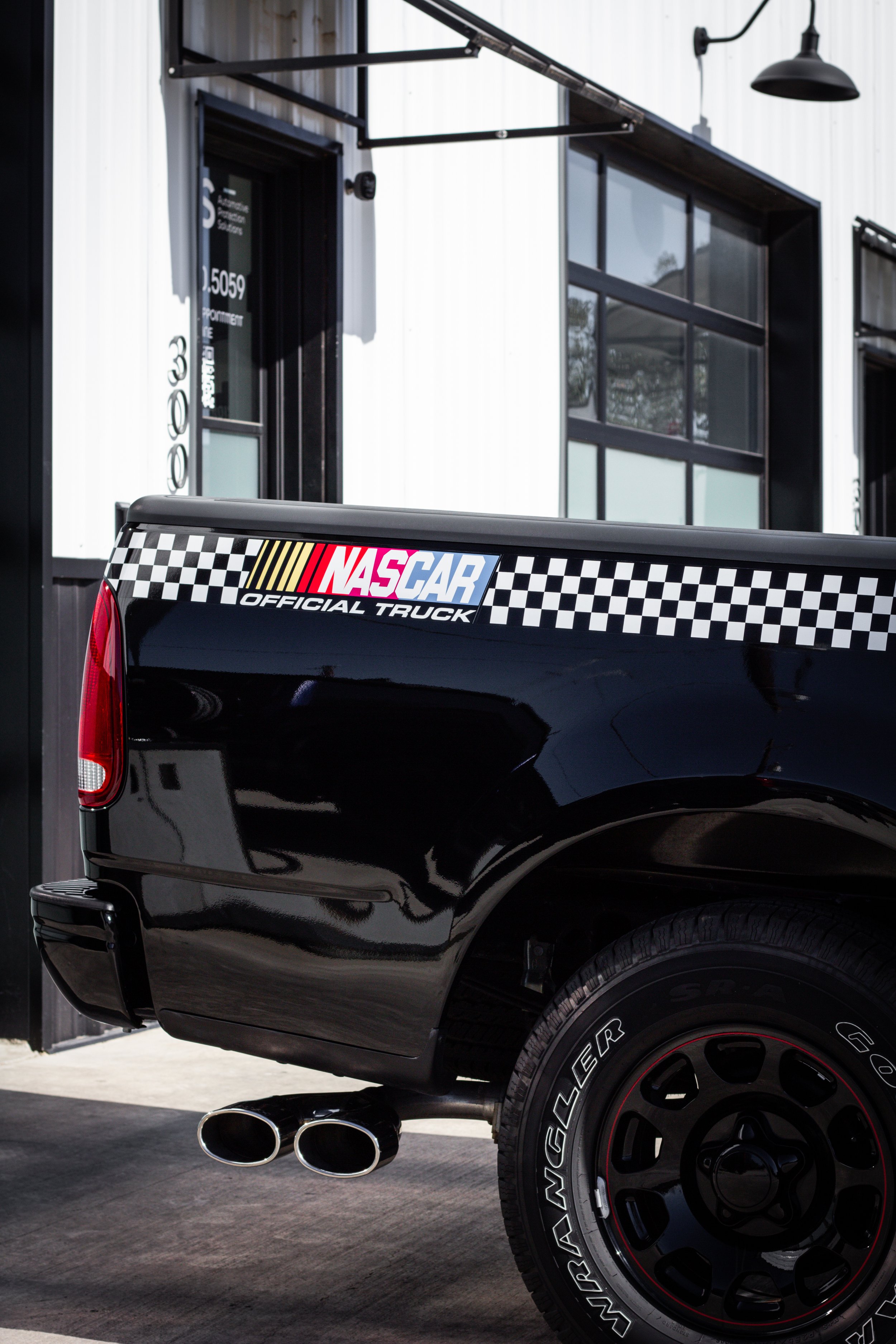 Camioneta negra con calcomanía de NASCAR y logotipo de truck oficial, estacionada frente a una edificación moderna con ventanas de vidrio y puertas negras.