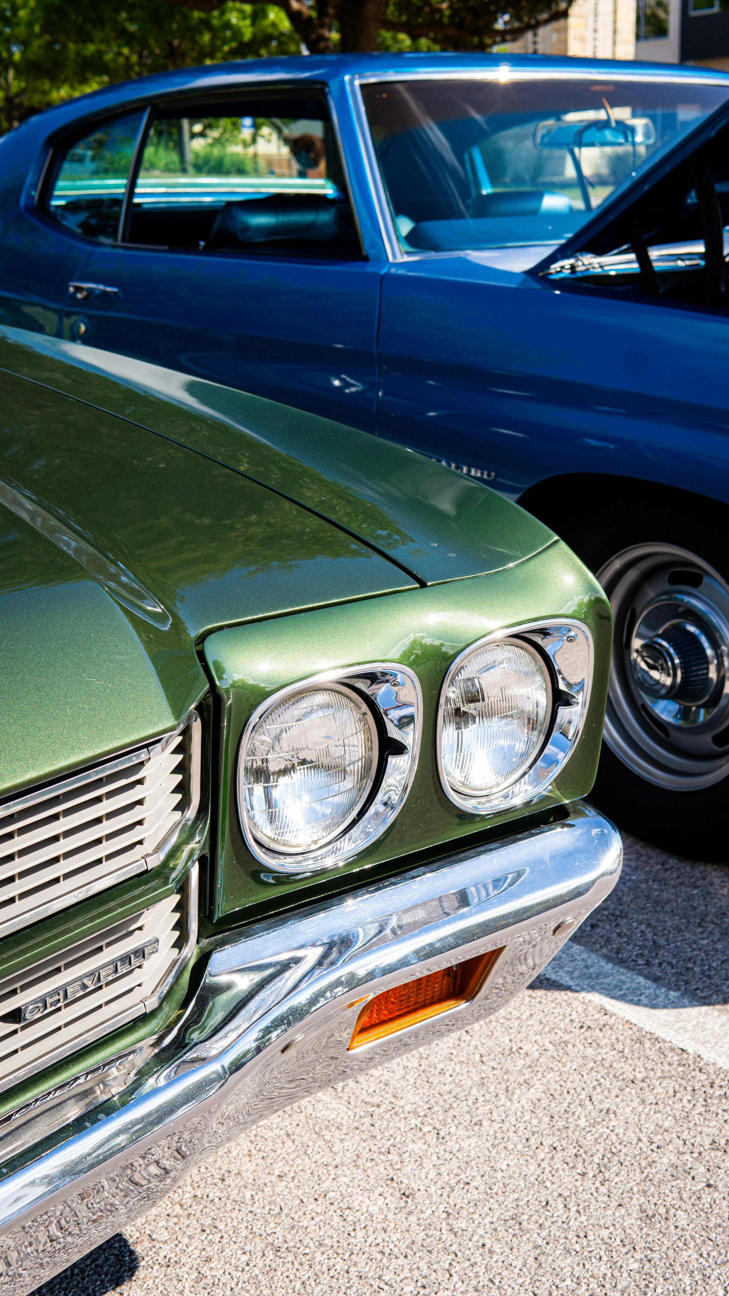 Primer plano del frente de dos autos clásicos, uno verde y uno azul, estacionados en una calle con árboles y edificios en el fondo, en un día soleado.