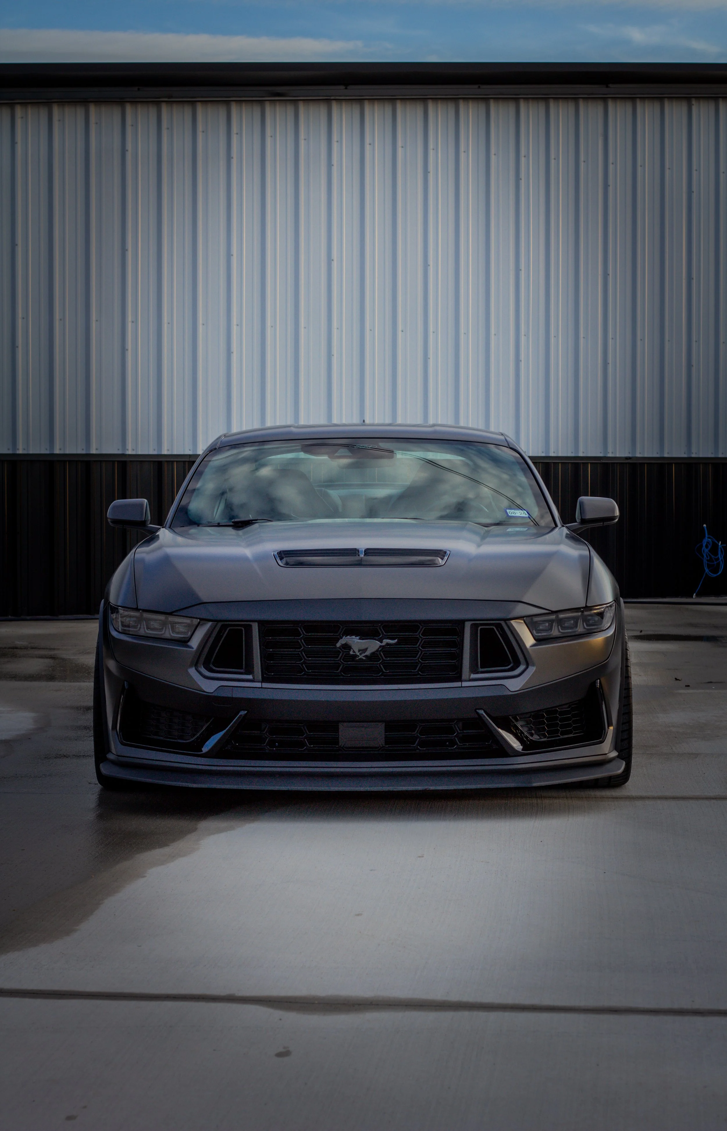 Auto deportivo Ford Mustang negro estacionado en un espacio de lavado o garaje, con pared metálica y cielo visible en el fondo.