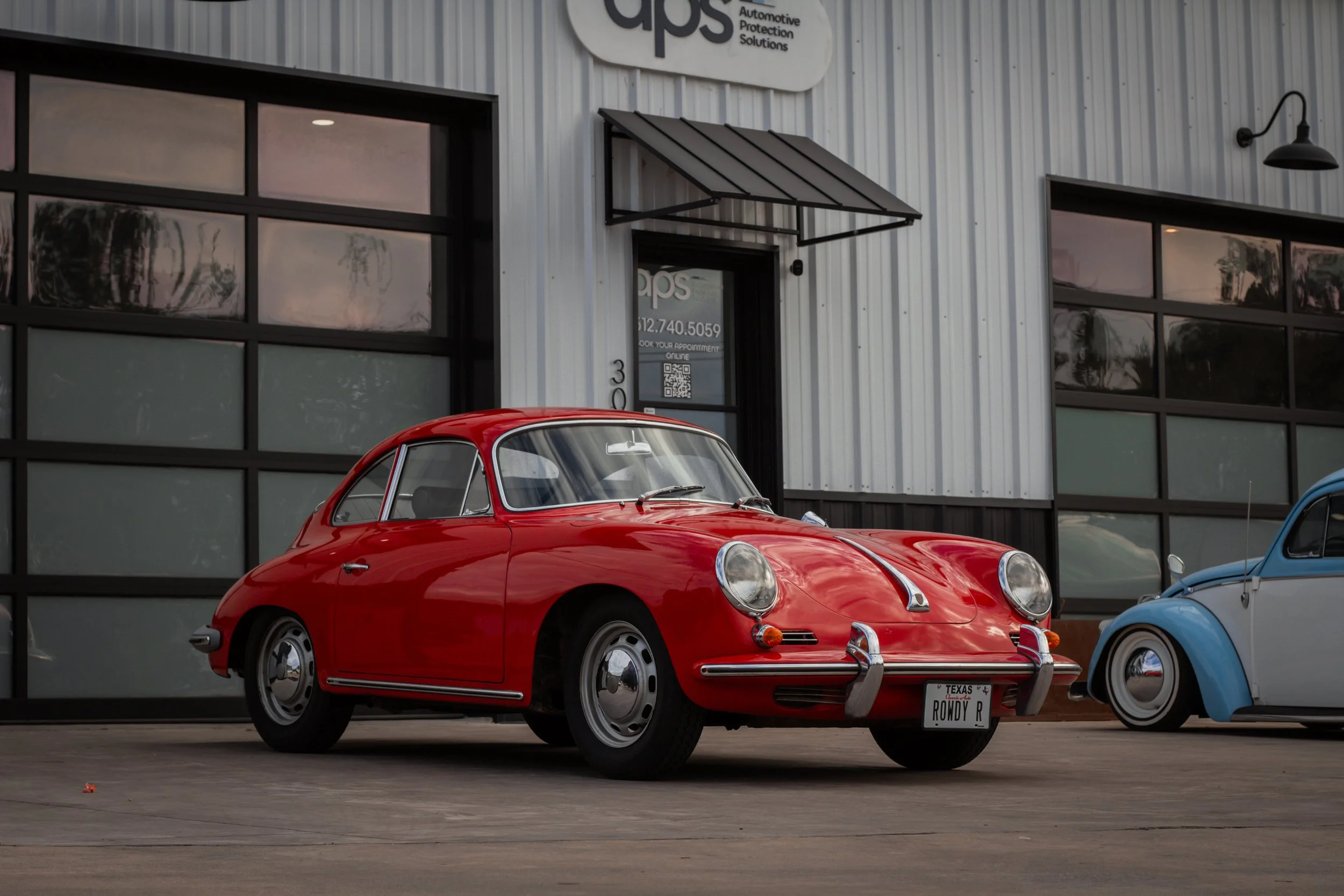 Coche clásico rojo estacionado frente a un edificio de estilo industrial con ventanas grandes y letrero que dice 'GPS' y 'Automotive Protection Solutions'.