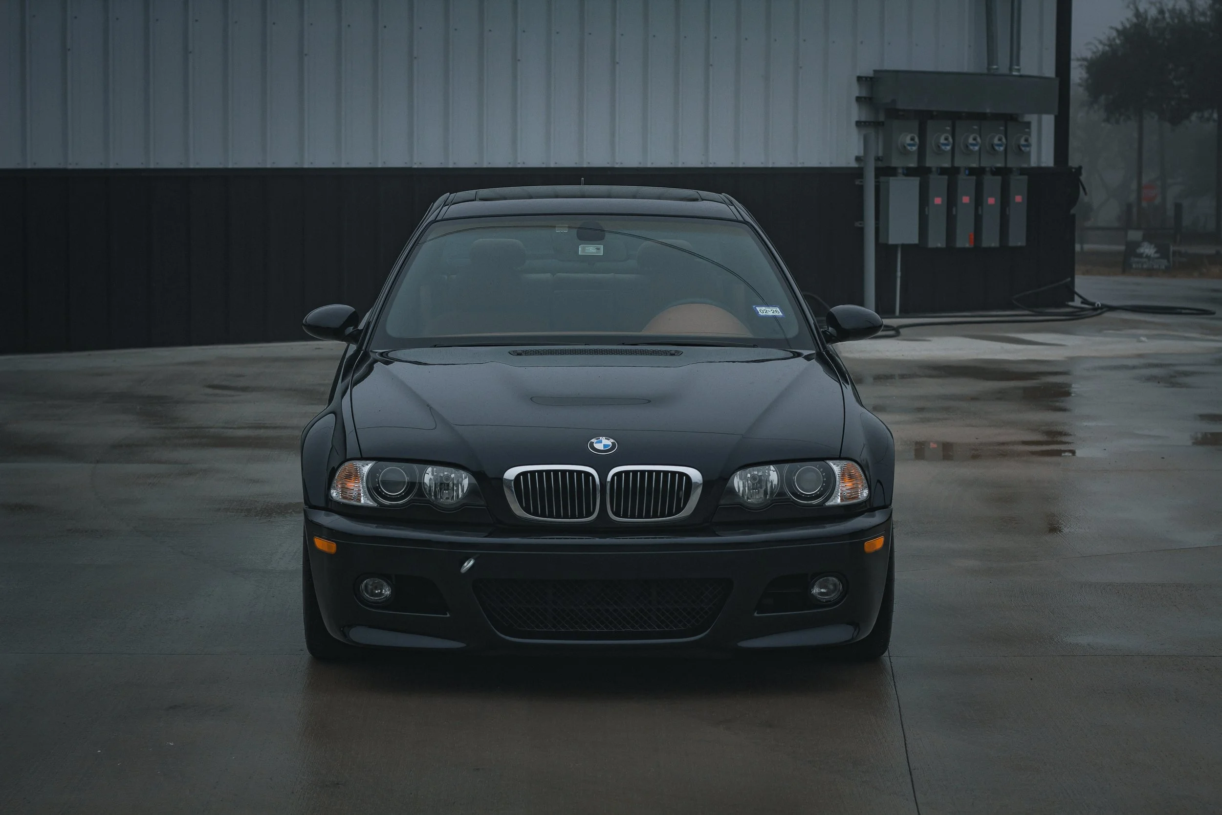 Coche negro tipo sedán BMW estacionado en un lugar con piso húmedo, edificio metálico de fondo y árboles al fondo.