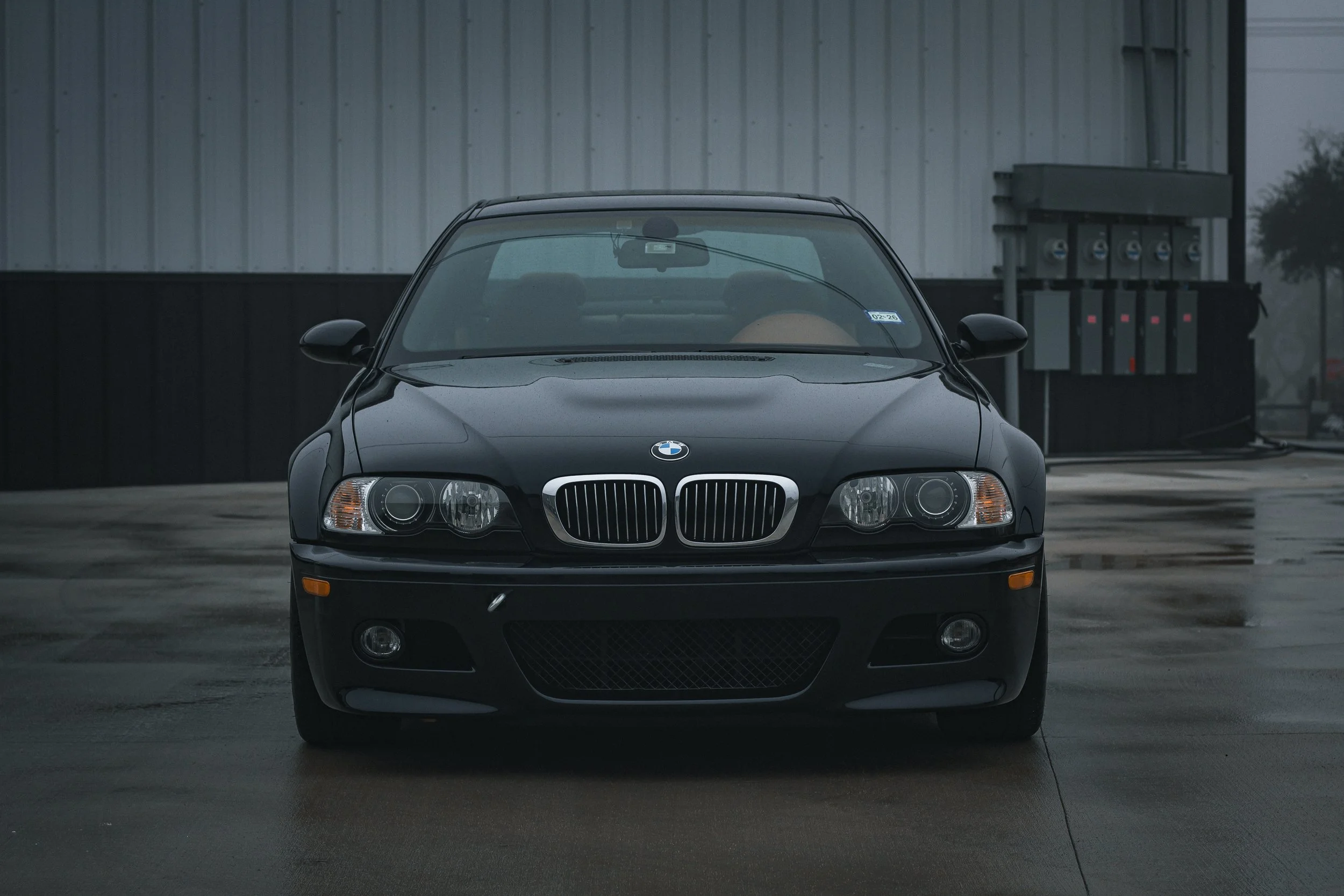 Coche BMW negro estacionado frente a pared de metal gris en un ambiente lluvioso.