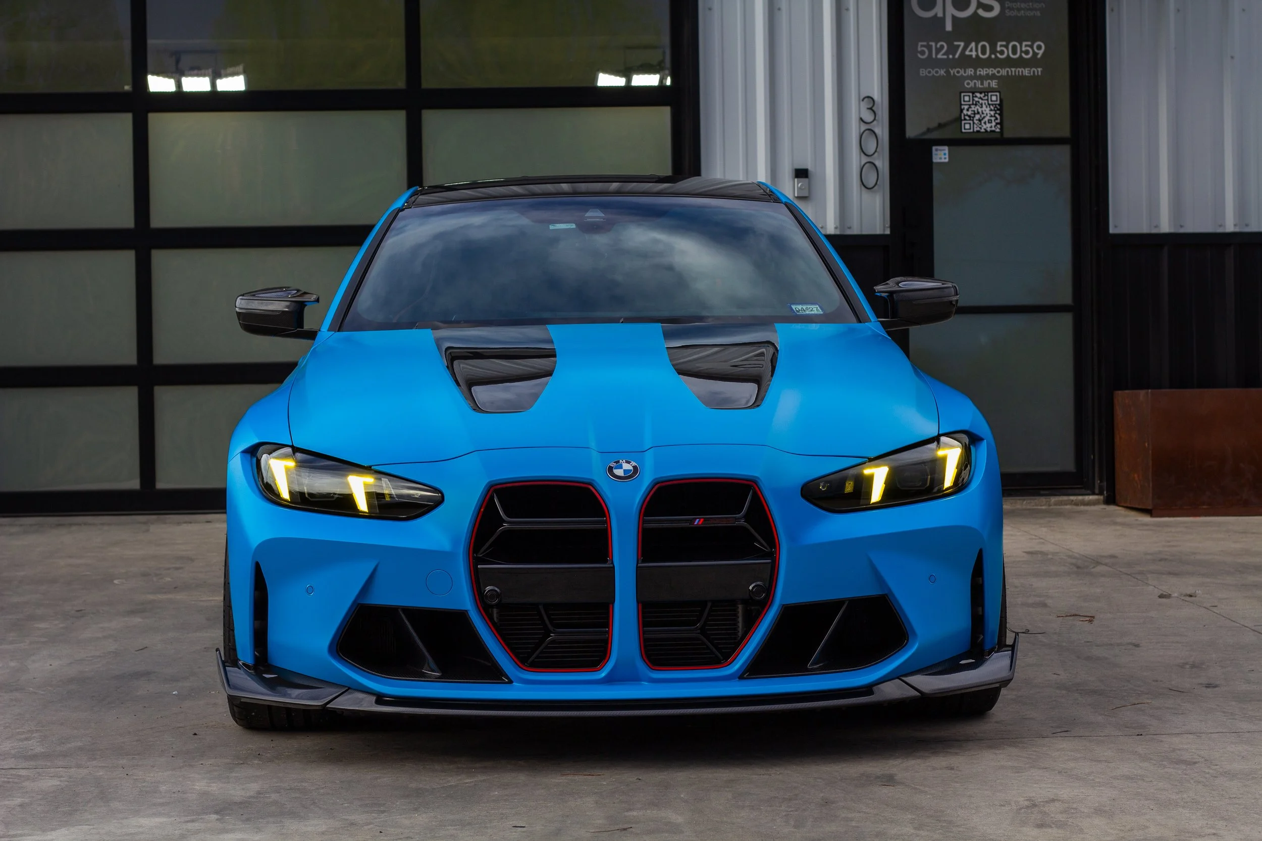 Un auto deportivo BMW azul con detalles en negro y rojo, estacionado frente a un edificio con ventanas y una puerta de entrada.