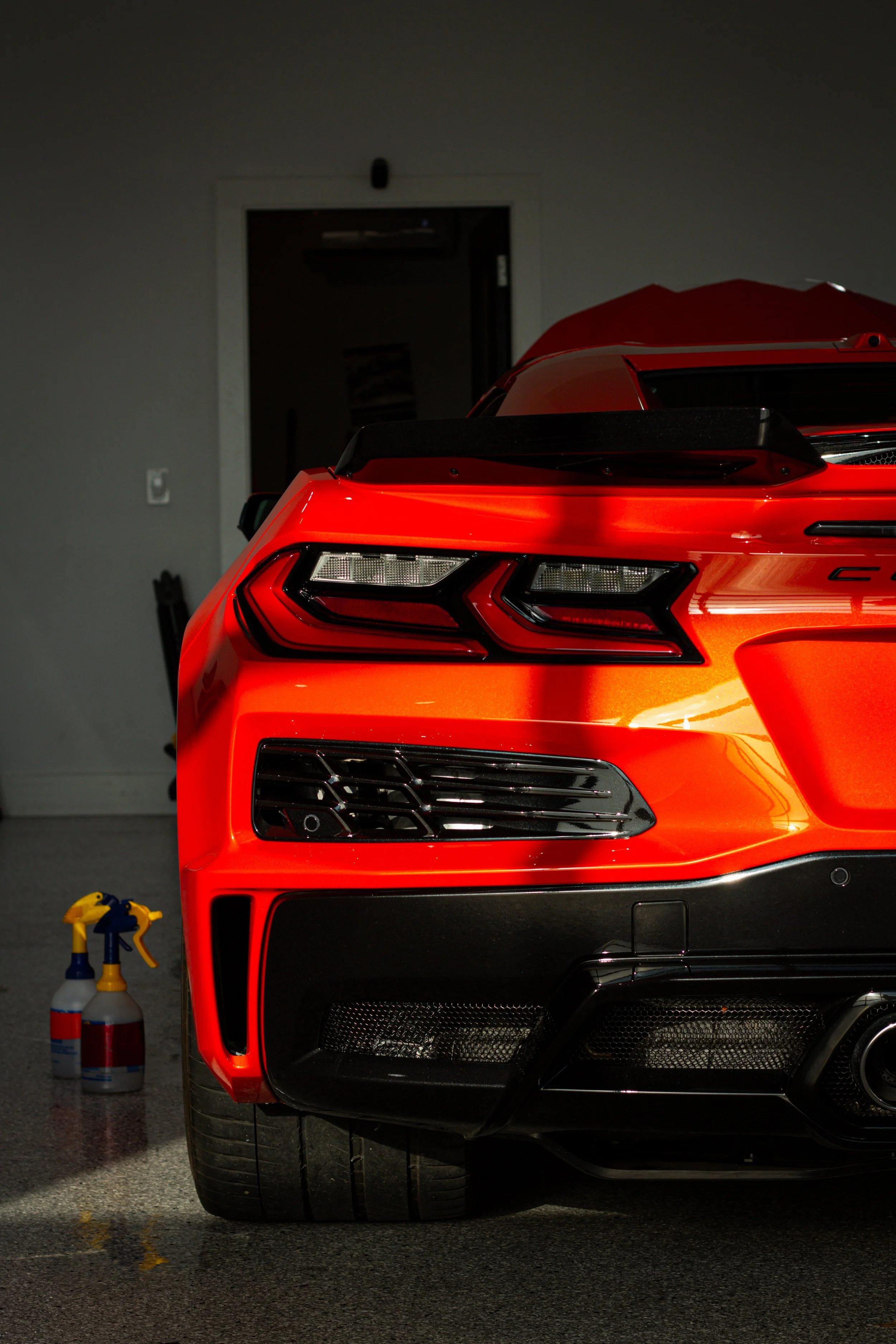 Parte trasera de un coche deportivo rojo en un garaje, con botellas rociadoras en el suelo a la izquierda.