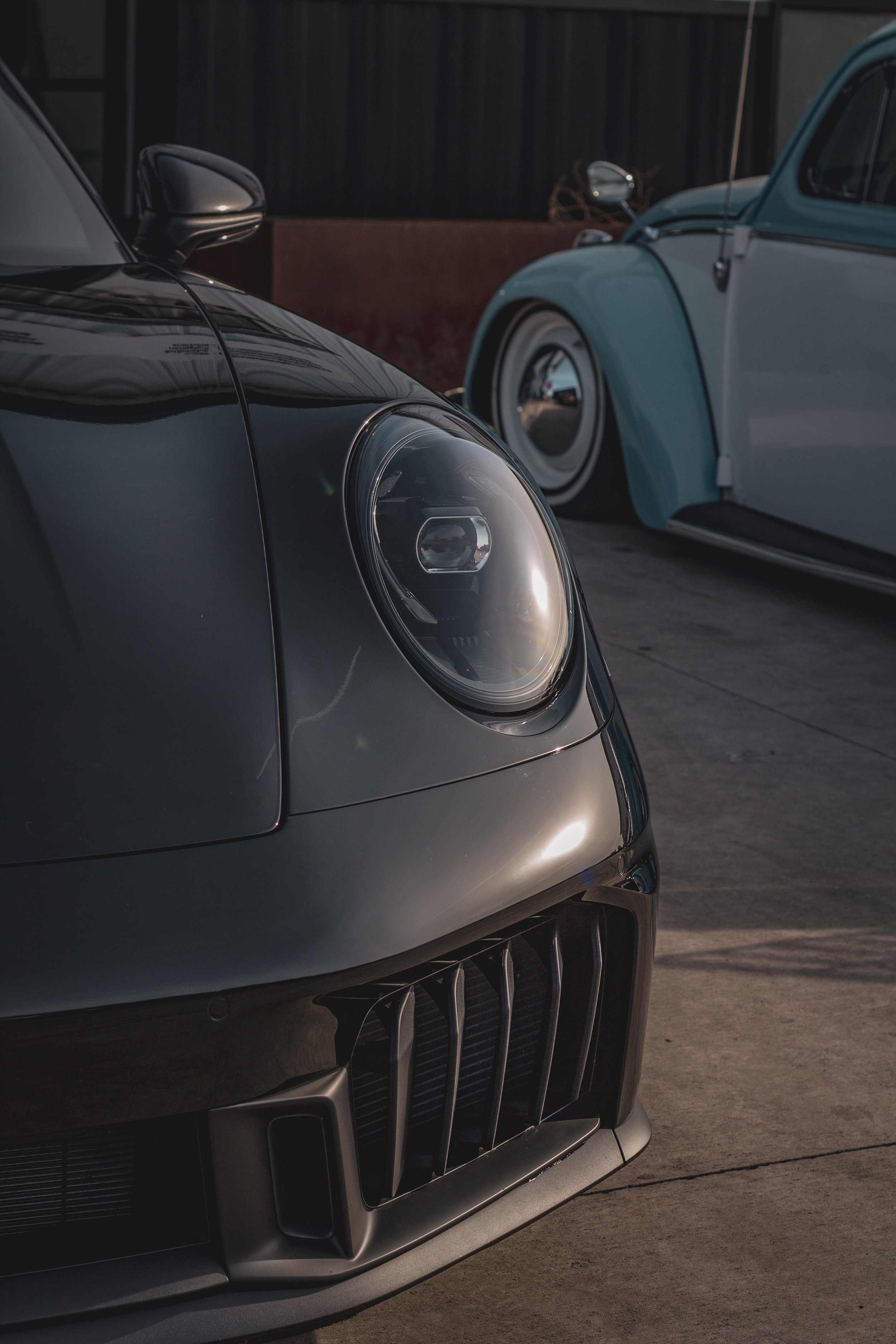Primer plano del coche deportivo negro con luces grandes y parrilla en la parte delantera, junto a otro coche clásico azul y blanco en el fondo.