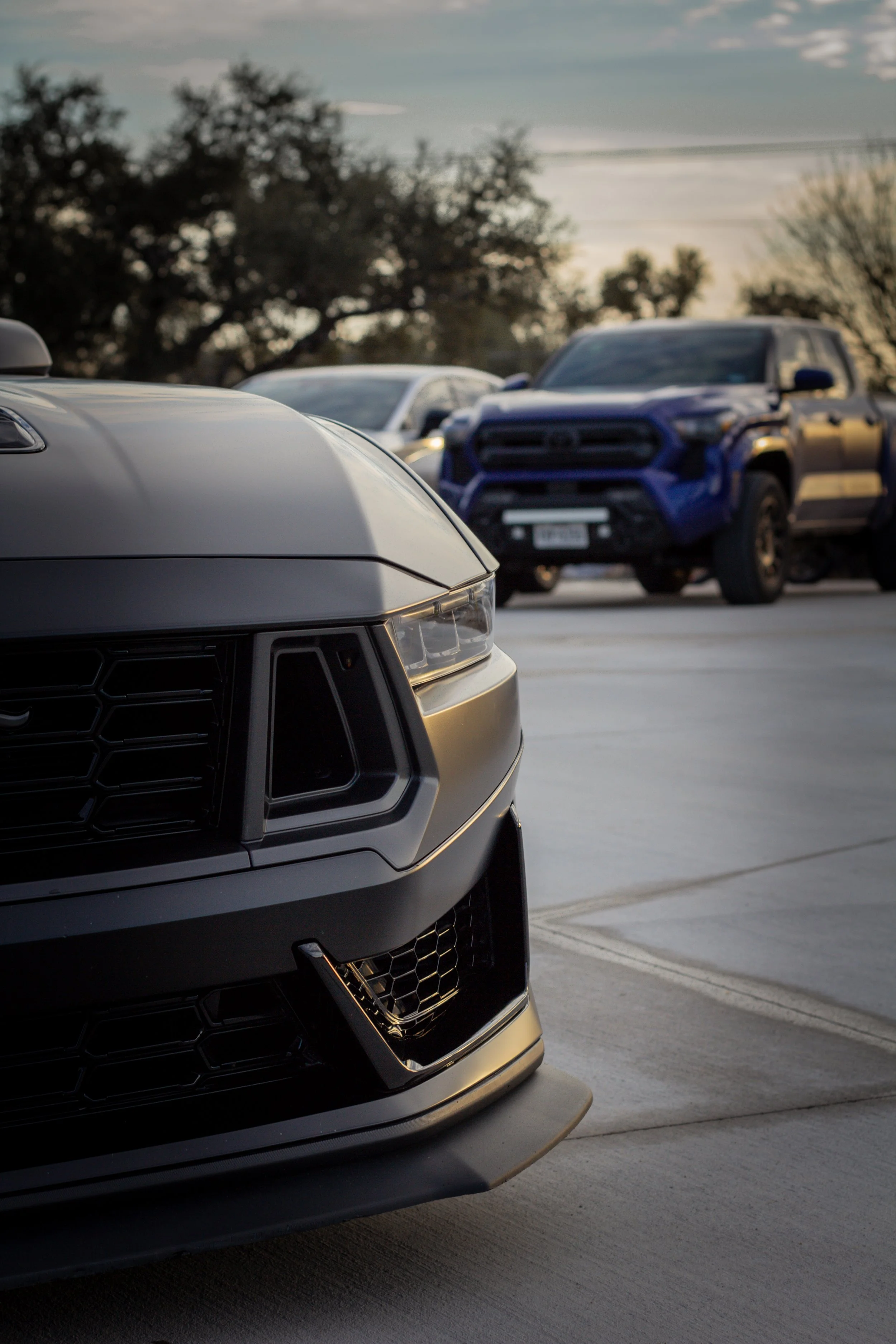 Primer plano de la parte frontal de un vehículo negro con faros y rejillas, con otros autos y árboles de fondo en un estacionamiento al atardecer.