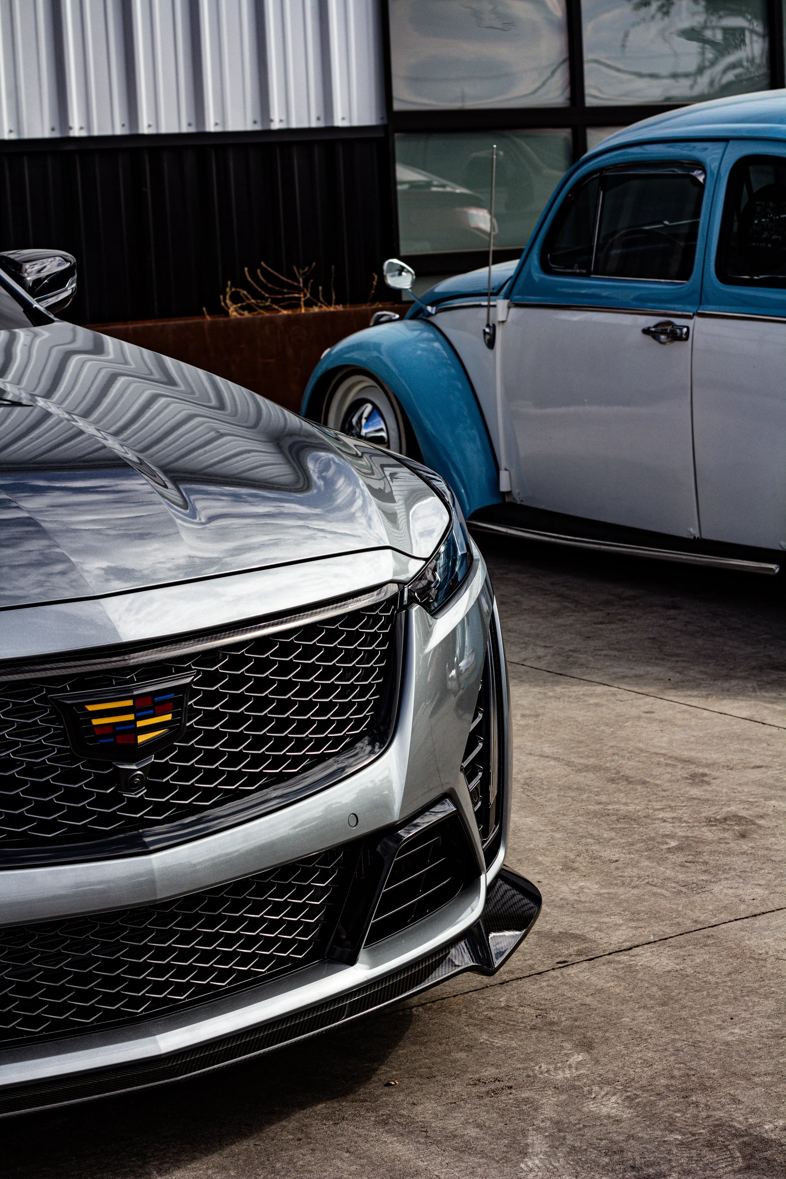 Un automóvil deportivo de color gris delante de un coche clásico azul y gris en un estacionamiento.