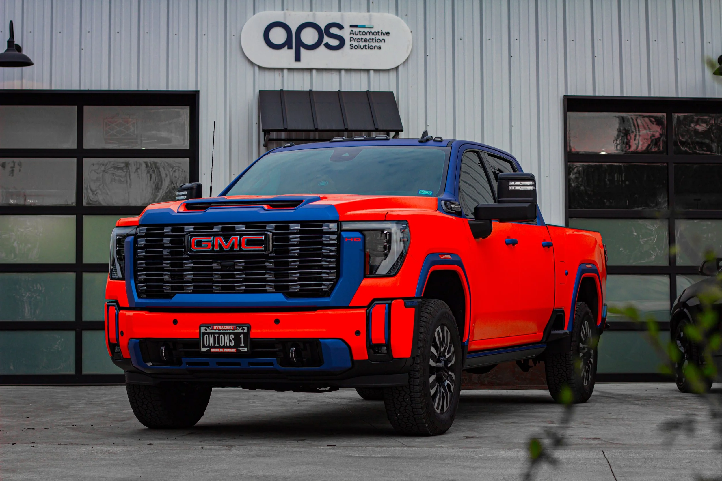Camioneta GMC de color naranja y azul estacionada frente a un edificio con logotipo de 'aps'.