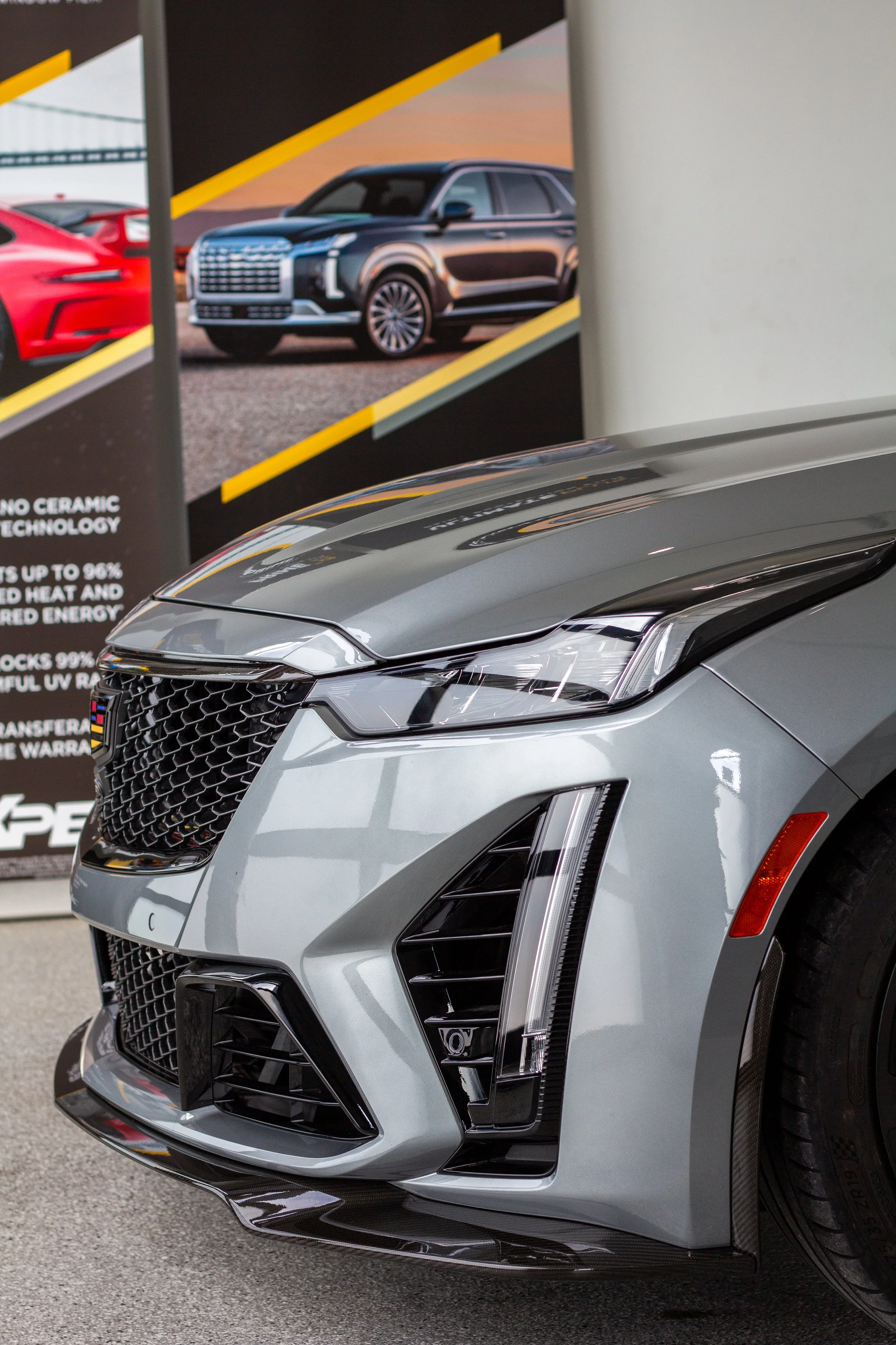 Primer plano del frente de un automóvil sedán de color gris en exhibición, con cartel promocional de autos y una imagen de otros vehículos en el fondo.