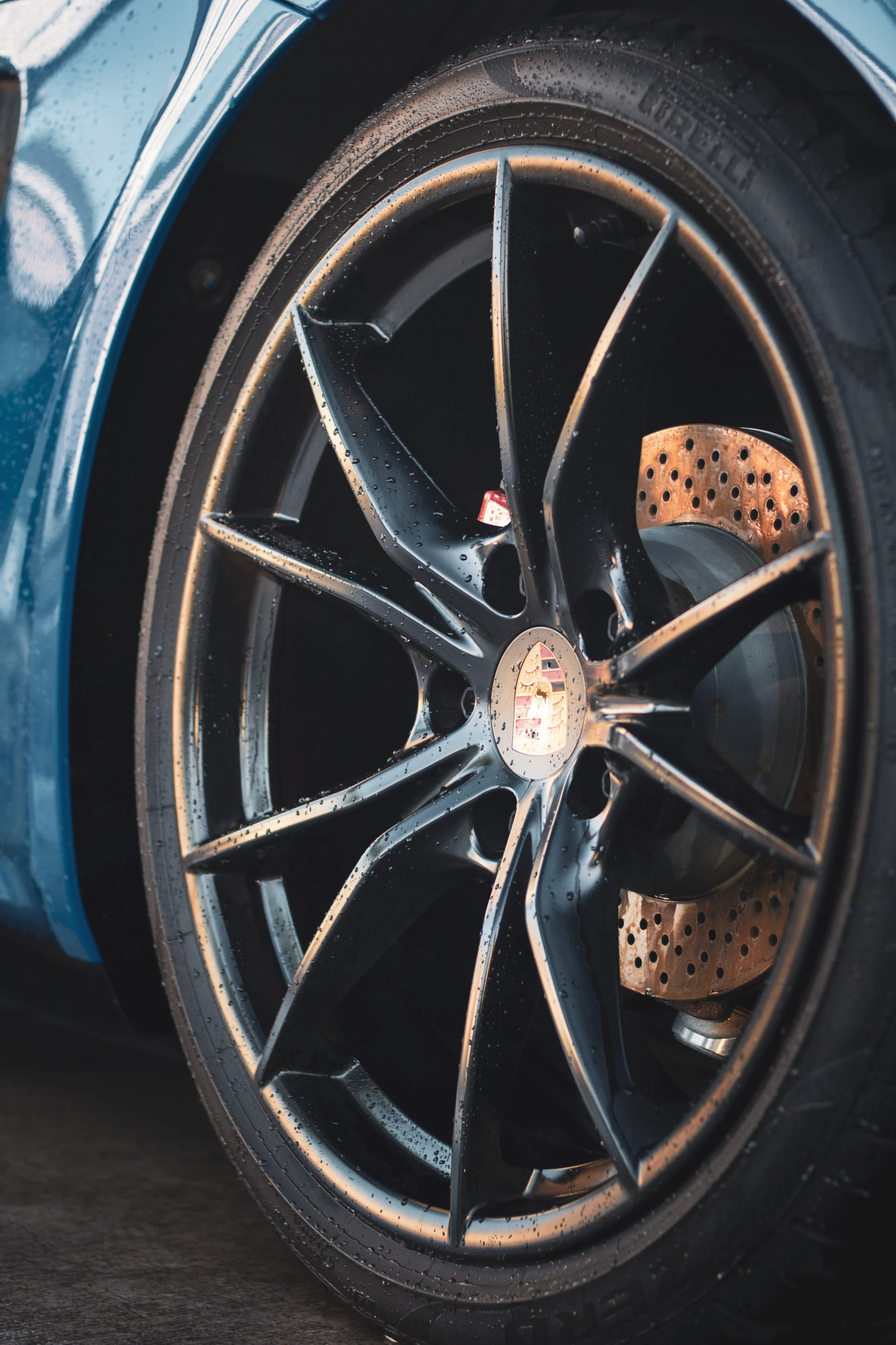 Rueda de un automóvil deportivo Porsche, con llanta negra y freno de disco perforado, en un coche azul con agua en la superficie.