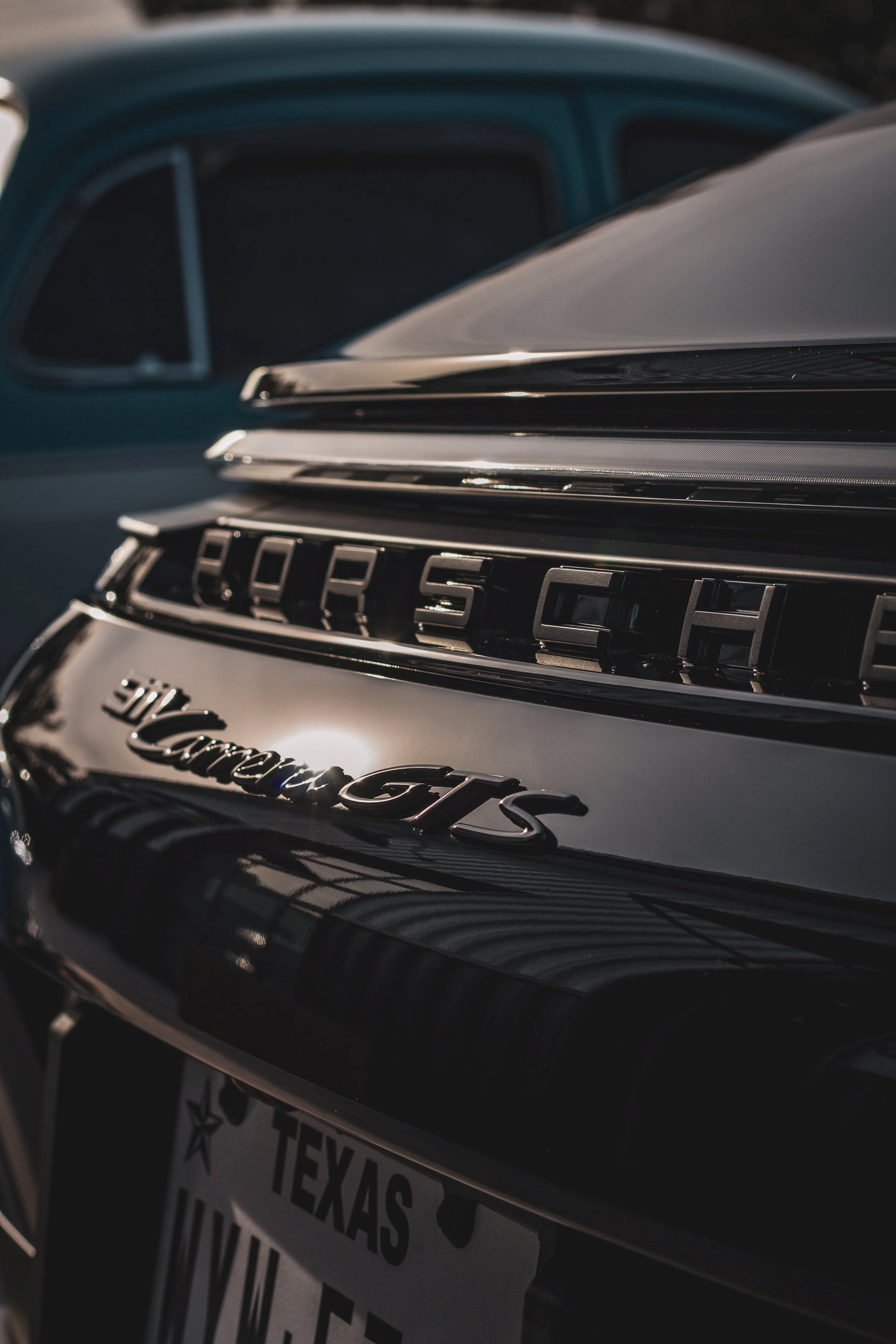 Cerca de la parte trasera de un auto Porsche con insignia Carrera GTS, con placas de Texas, y otro coche azul al fondo.