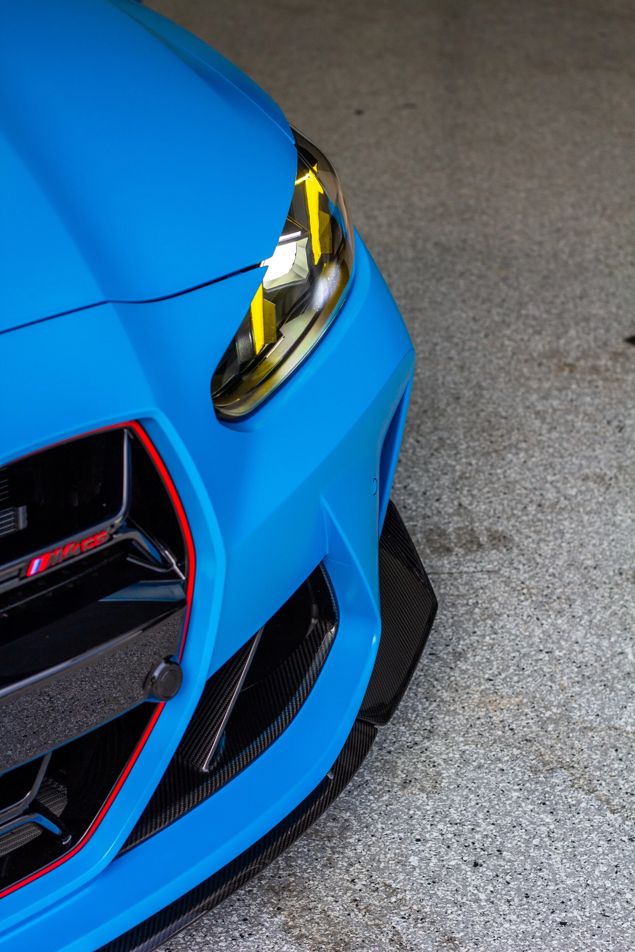 Primer plano de la parte delantera de un coche deportivo azul, mostrando el faro y el parachoques con detalles en fibra de carbono.