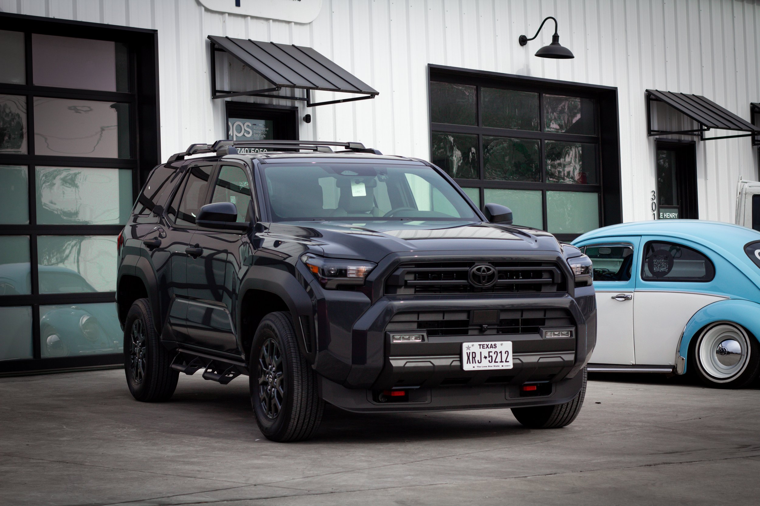 Camioneta Toyota negra y coche clásico azul y blanco estacionados en un garaje frente a un edificio con ventanas y puertas de cristal.