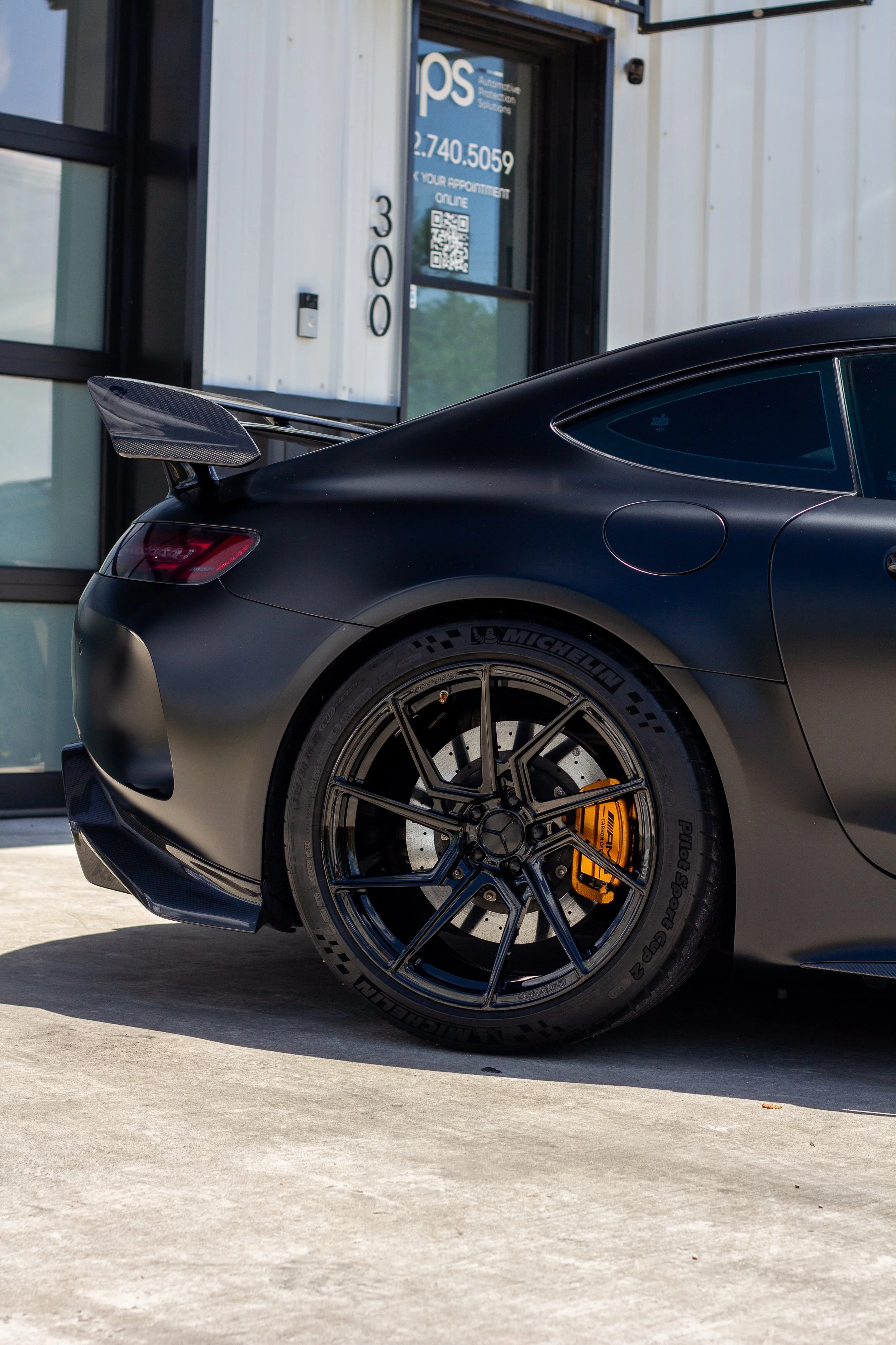 Parte trasera de un automóvil deportivo negro con llantas Michelin y frenos de colores amarillo, estacionado frente a un edificio con cartel que dice 'PS' y un número de teléfono.