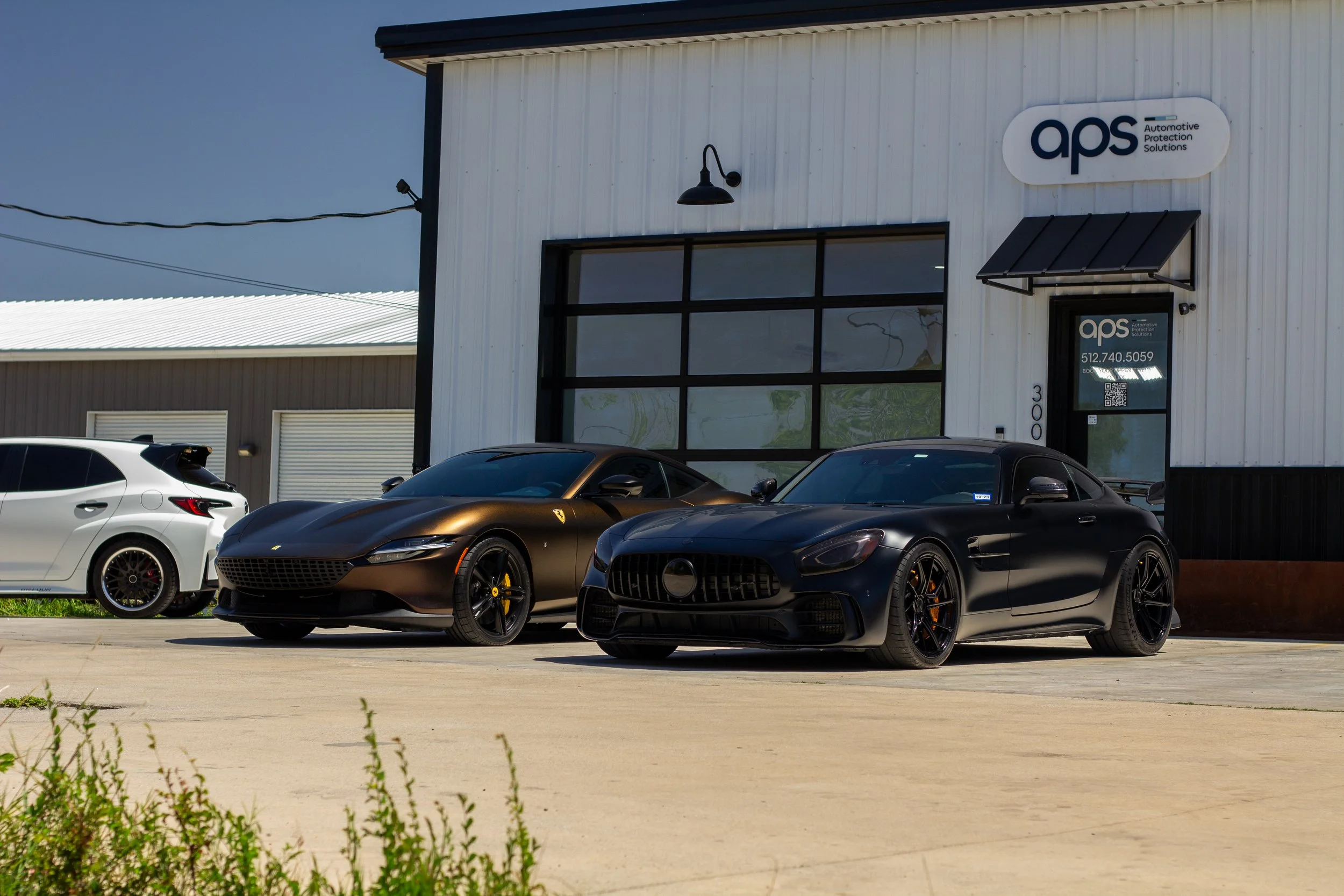 Tres autos de lujo, incluyendo un Ferrari negro, un Mercedes-AMG negro y un auto blanco, estacionados frente a un taller de reparación de autos con el letrero APS.