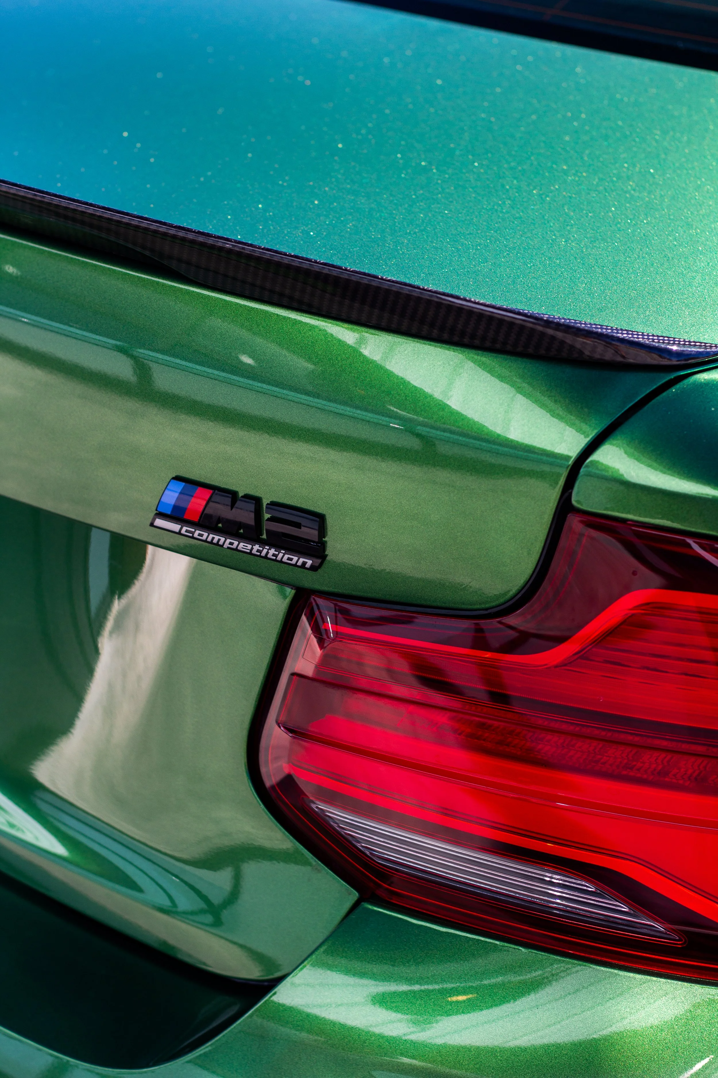 Imagen de la parte trasera de un coche verde con un emblema 'M' y la palabra 'competition', y una luz trasera roja.