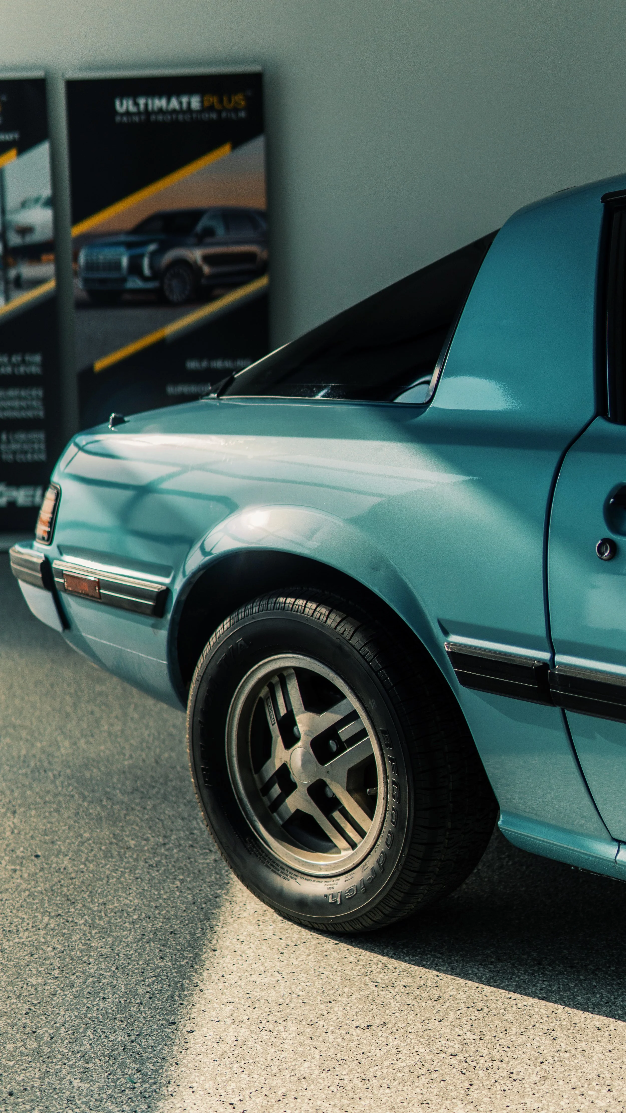 Parte frontal de un coche clásico azul, mostrando la rueda y la puerta, con cartel de promoción de autos en el fondo.