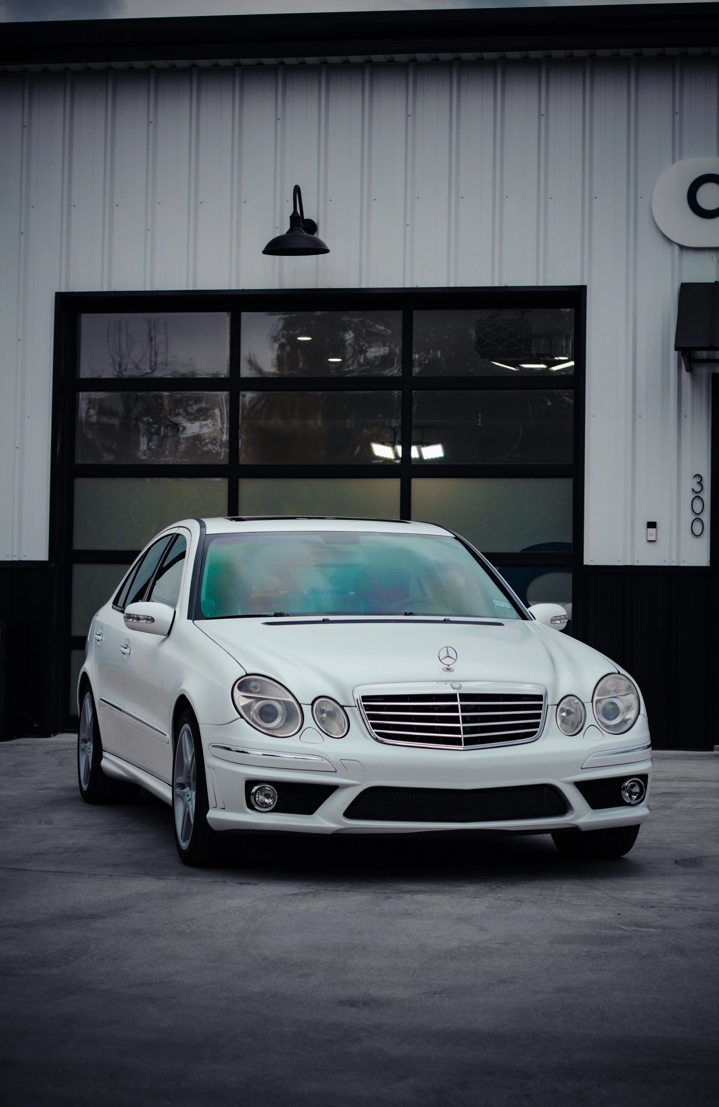 Auto sedán Mercedes-Benz blanco estacionado frente a un edificio de fondo con ventanas negras.