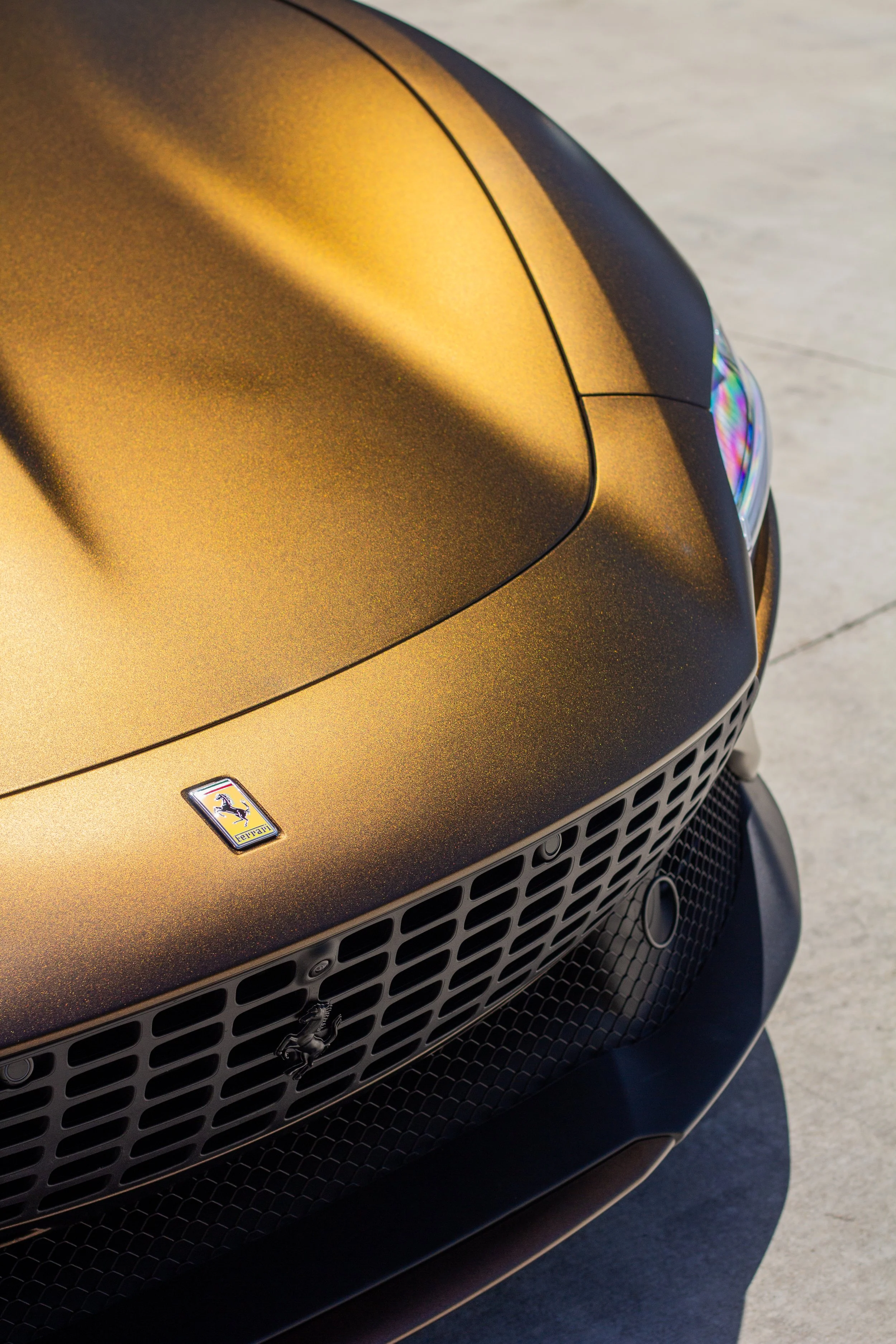 Primer plano de la parte delantera de un automóvil Ferrari de color dorado con detalles en negro, mostrando el logotipo de Ferrari en el capó y una parrilla delantera grande.