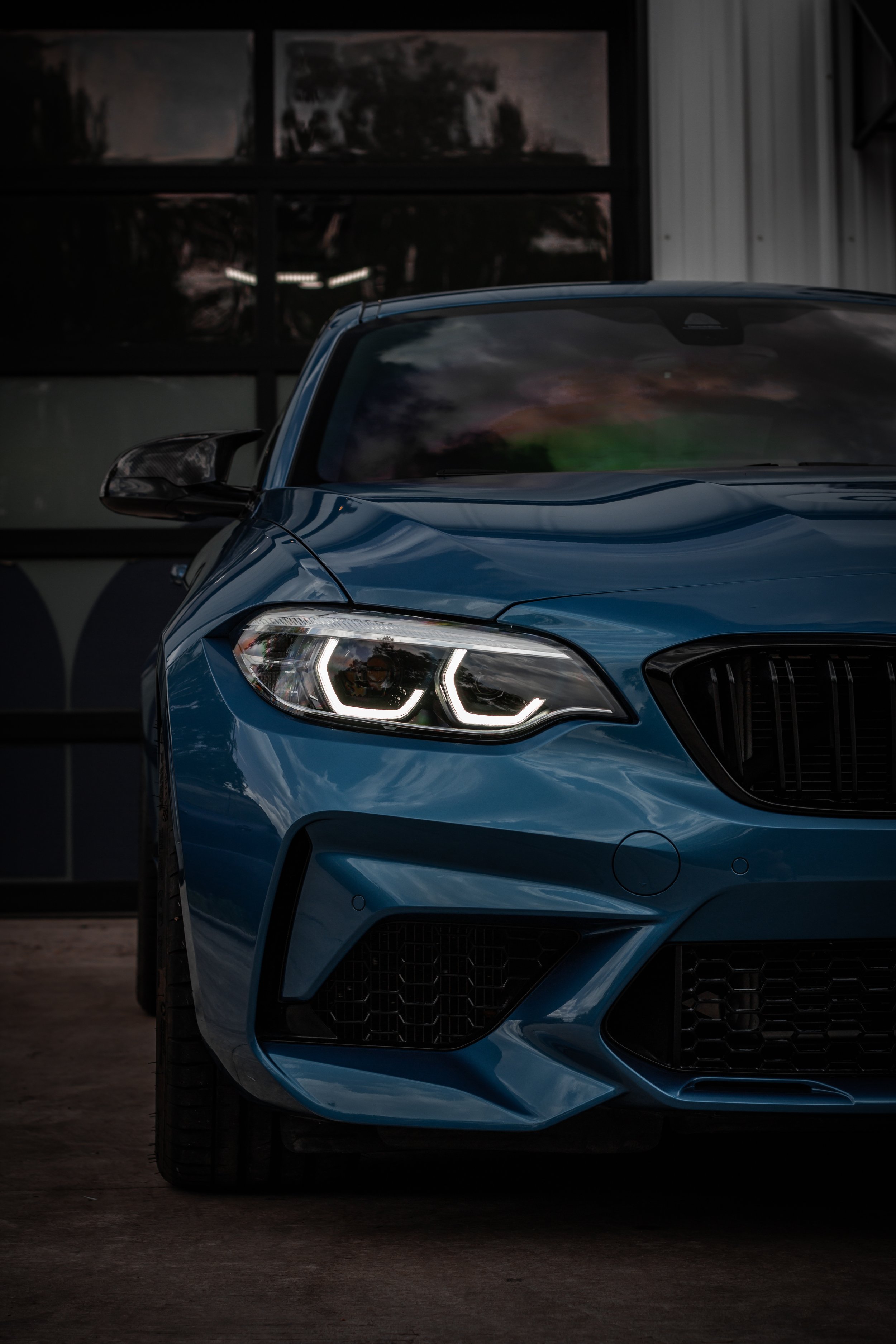 Frontal de un coche deportivo azul oscuro con faros LED y parrilla negra, estacionado en un garaje con paredes blancas y ventanas oscuras.