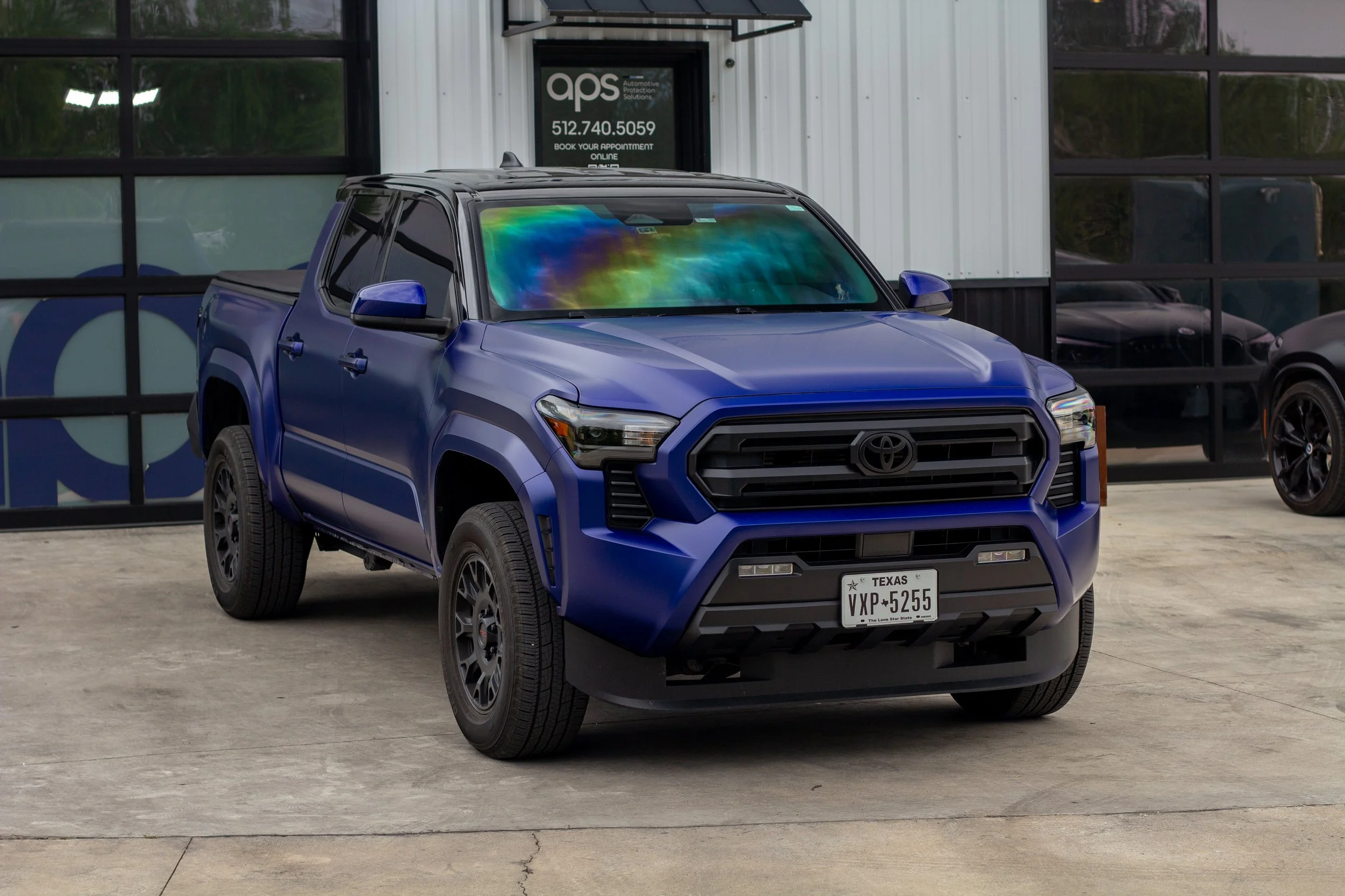 Camioneta Toyota azul estacionada frente a una tienda de autopartes con ventanas grandes y un letrero que dice 'APS' y 'Reserva tu cita en línea'.