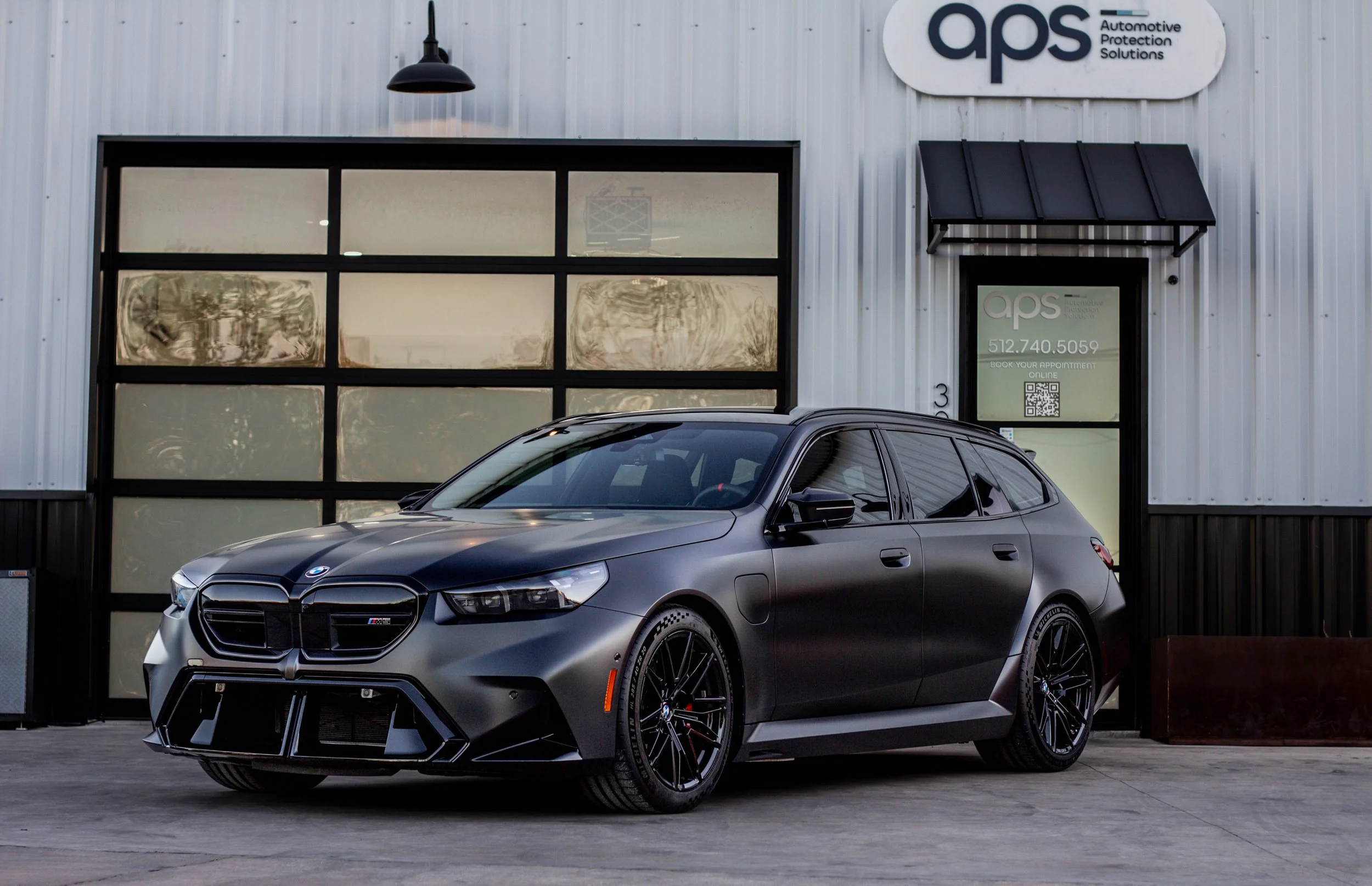 Automóvil BMW serie 3 en color gris oscuro estacionado frente a un edificio con logotipo 'aps'.