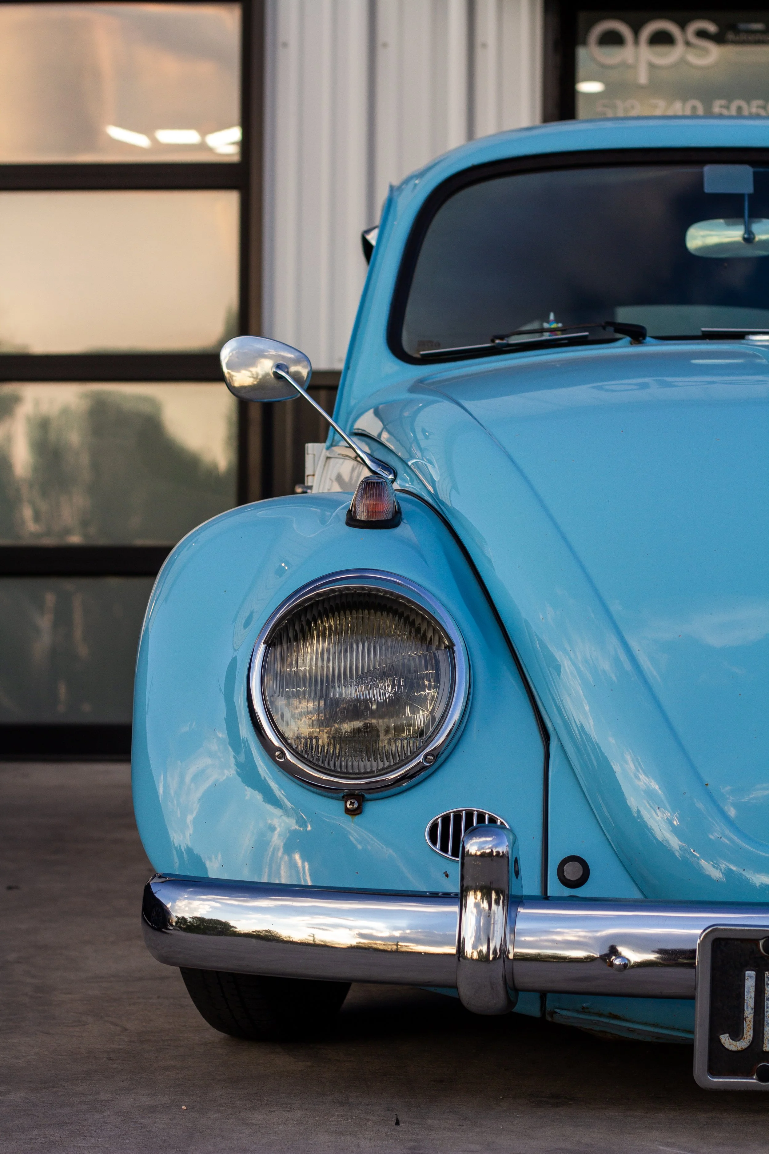Parte delantera de un coche clásico de color azul, con faro, espejo retrovisor y parachoques cromados, estacionado frente a una estructura moderna.