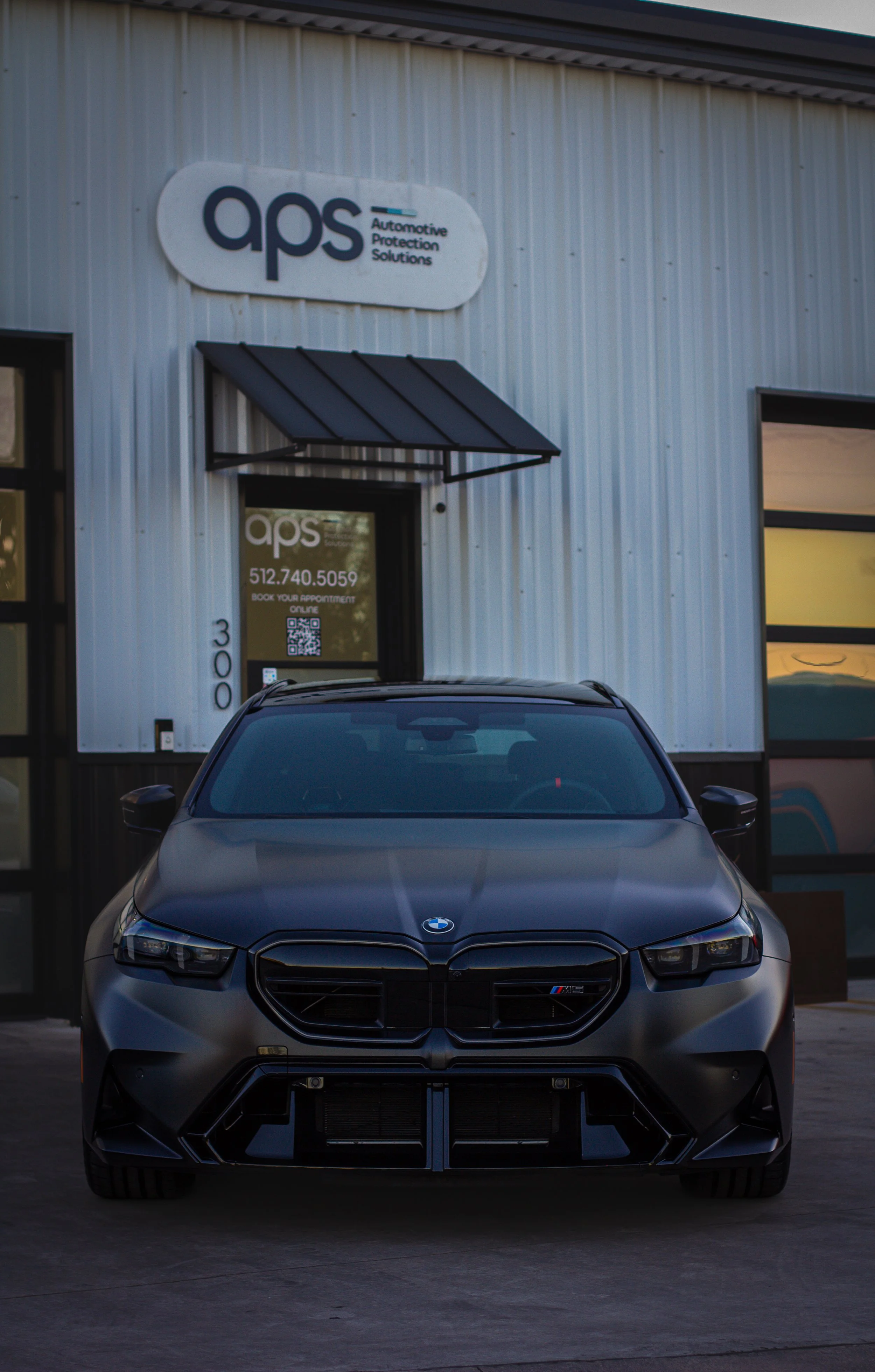 Coche negro BMW M3 estacionado frente a una clínica de protección automotriz llamada APS.