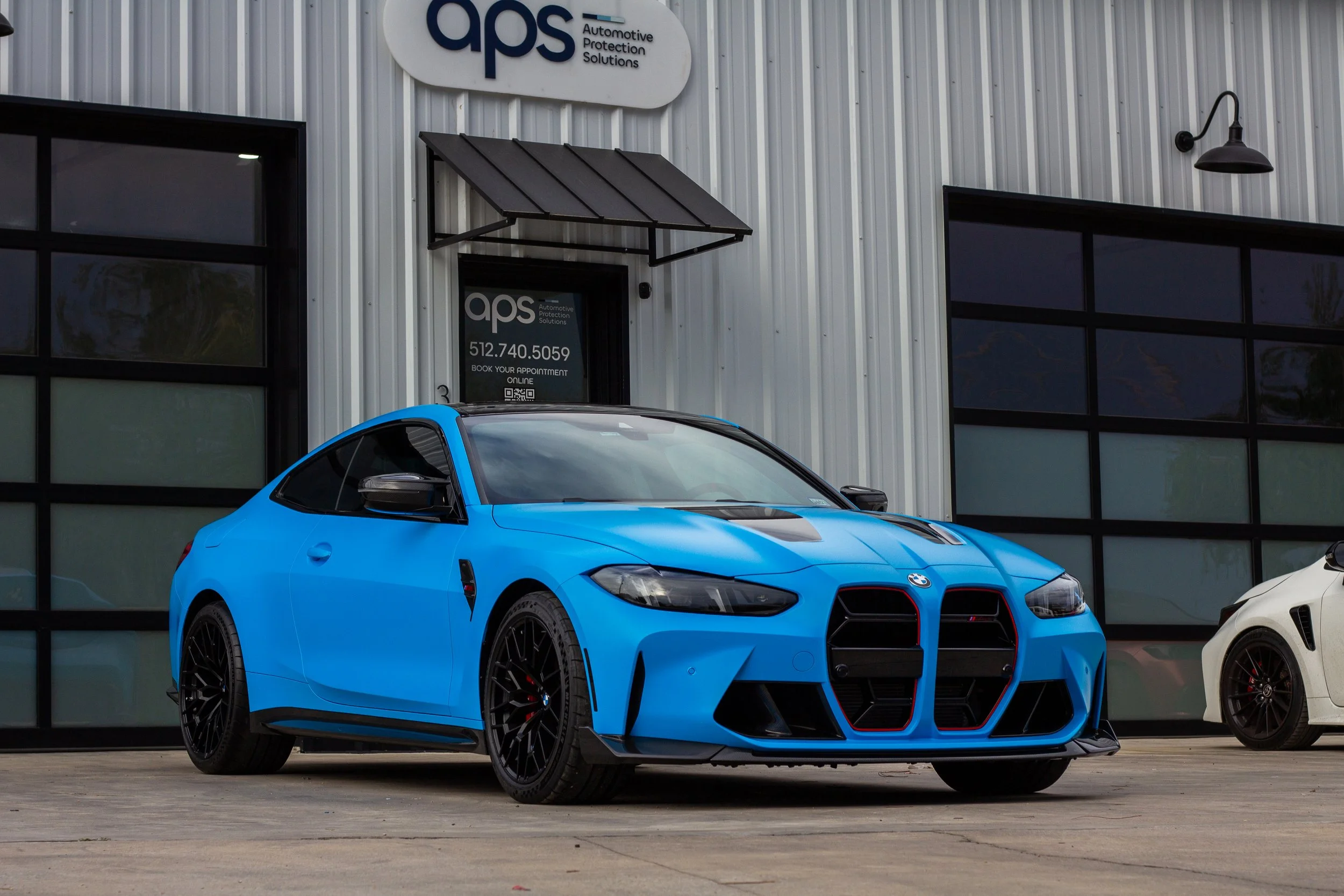 Auto deportivo azul con diseño aerodinámico en exhibición frente a un edificio con cartel de APS, en un área de estacionamiento.