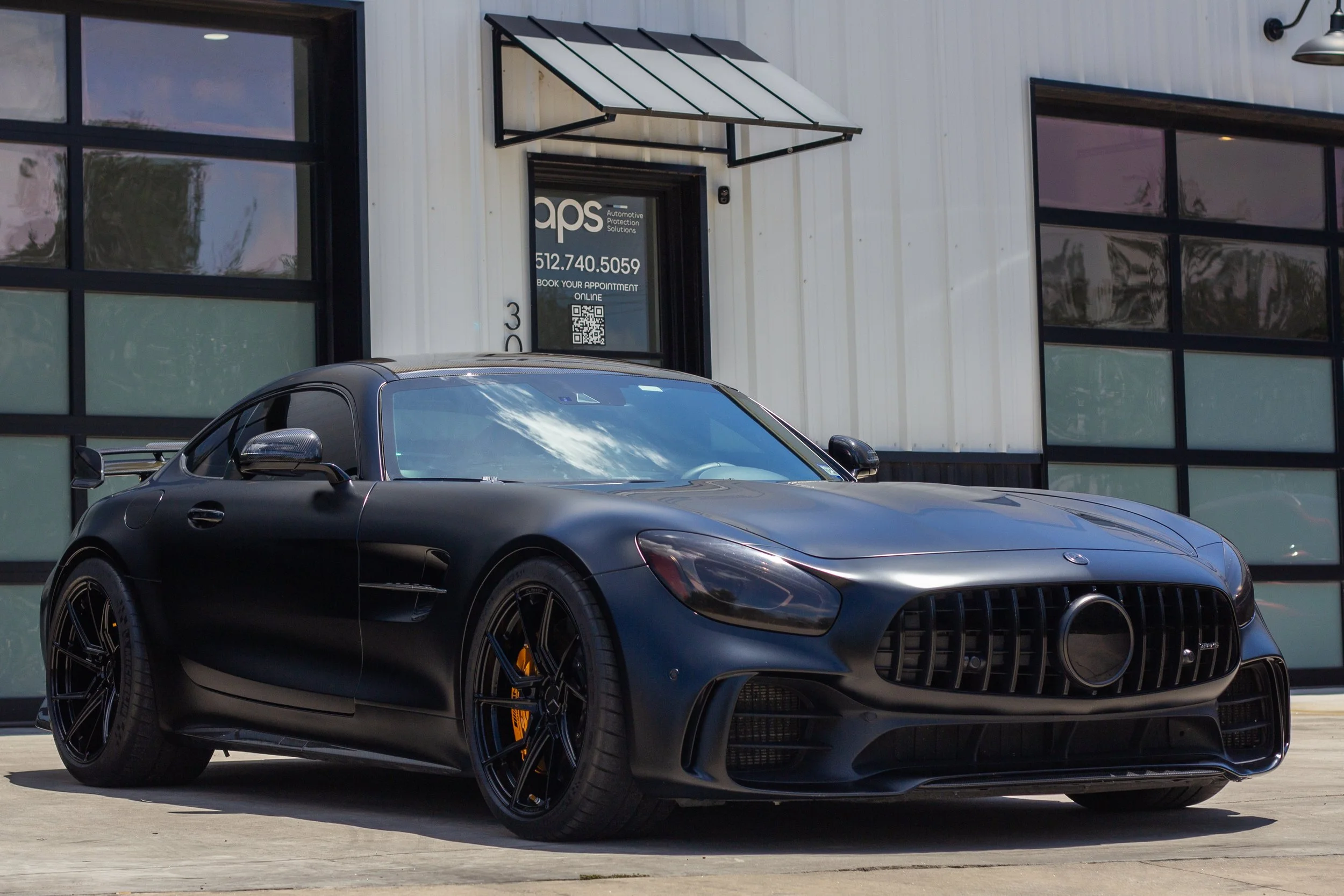 Coche deportivo negro con detalles en negro, estacionado frente a un edificio con ventanas grandes y una puerta de garaje, en un día soleado.
