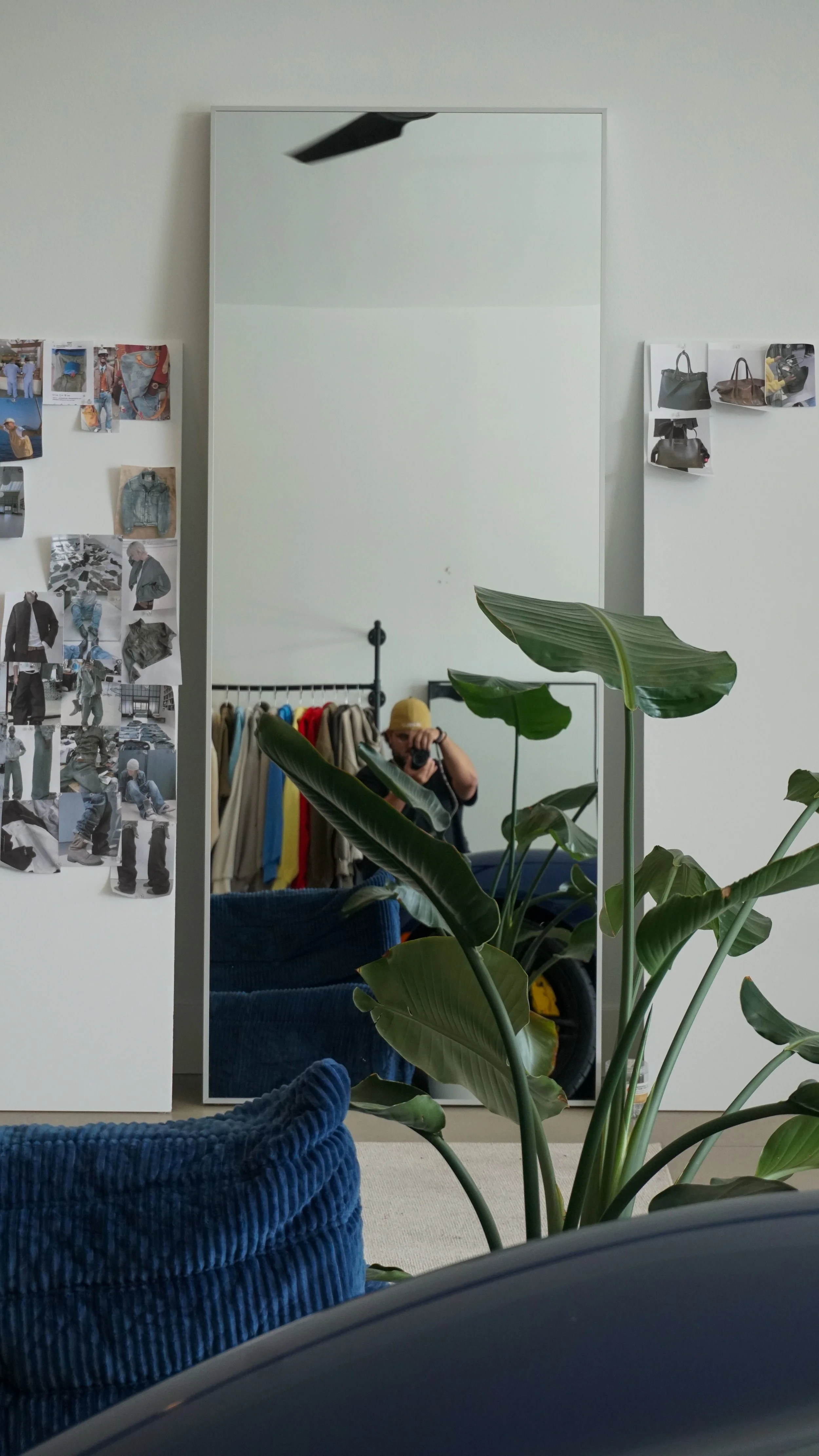Reflection en un espejo de una persona con gorra que se toma una selfie, rodeada de plantas y ropa colgada, en un espacio con paredes blancas con fotos y recortes pegados.