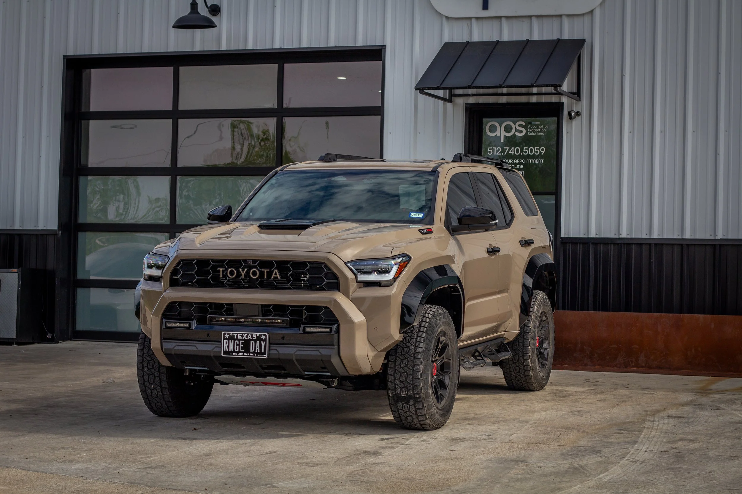 Vehículo todoterreno color beige estacionado frente a un edificio con ventanas y letrero. Modelo de Toyota con matrícula de Texas y neumáticos grandes.