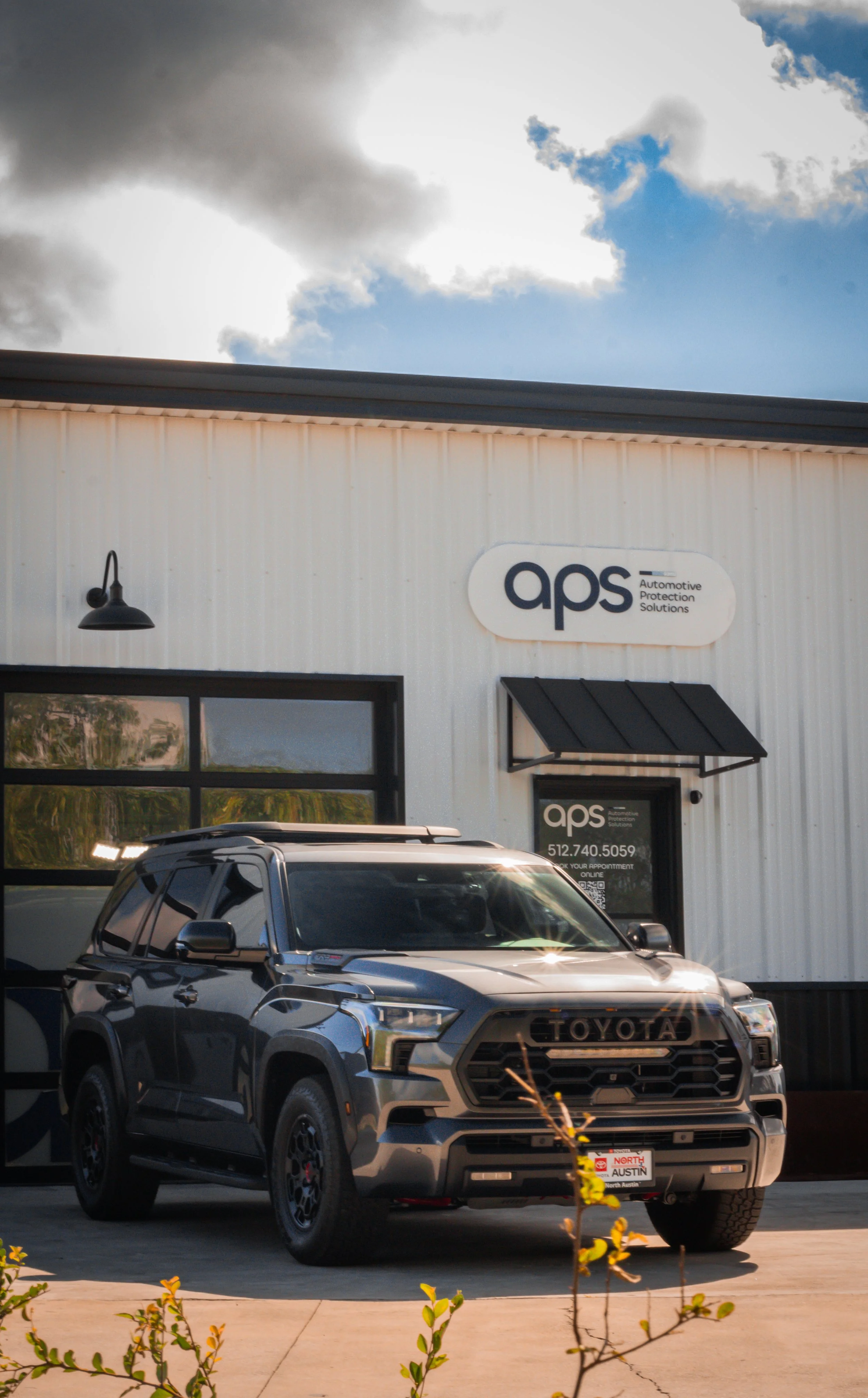 Frontal de un vehículo Toyota negro estacionado frente a un edificio de color blanco con un letrero que dice 'aps' y 'Automotive Protection Solutions', con un cielo parcialmente nublado en el fondo.