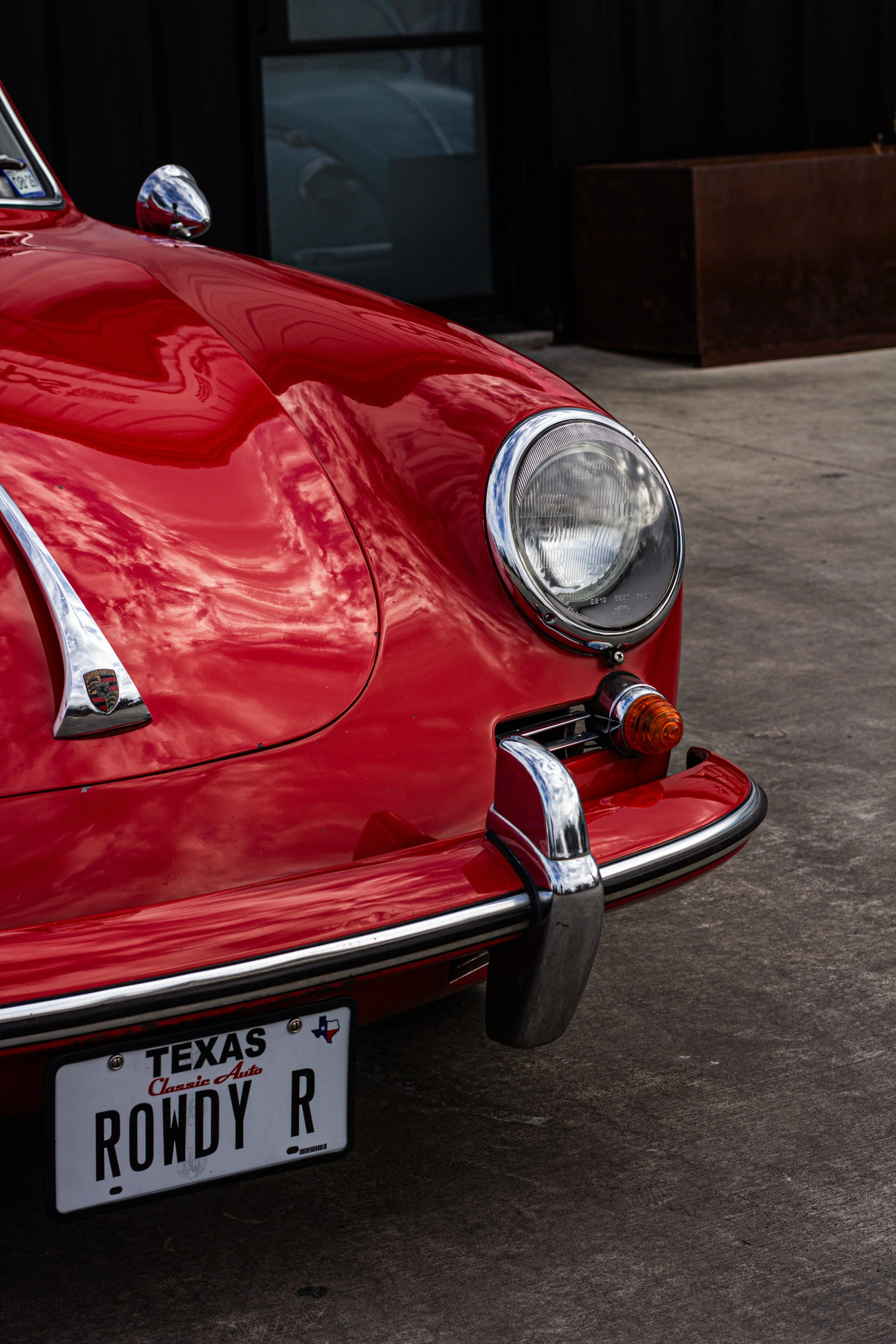 Frontal de un automóvil clásico rojo, prominently showing su faro, parrilla, y matrícula con el texto 'TEXAS ROWDY R'.