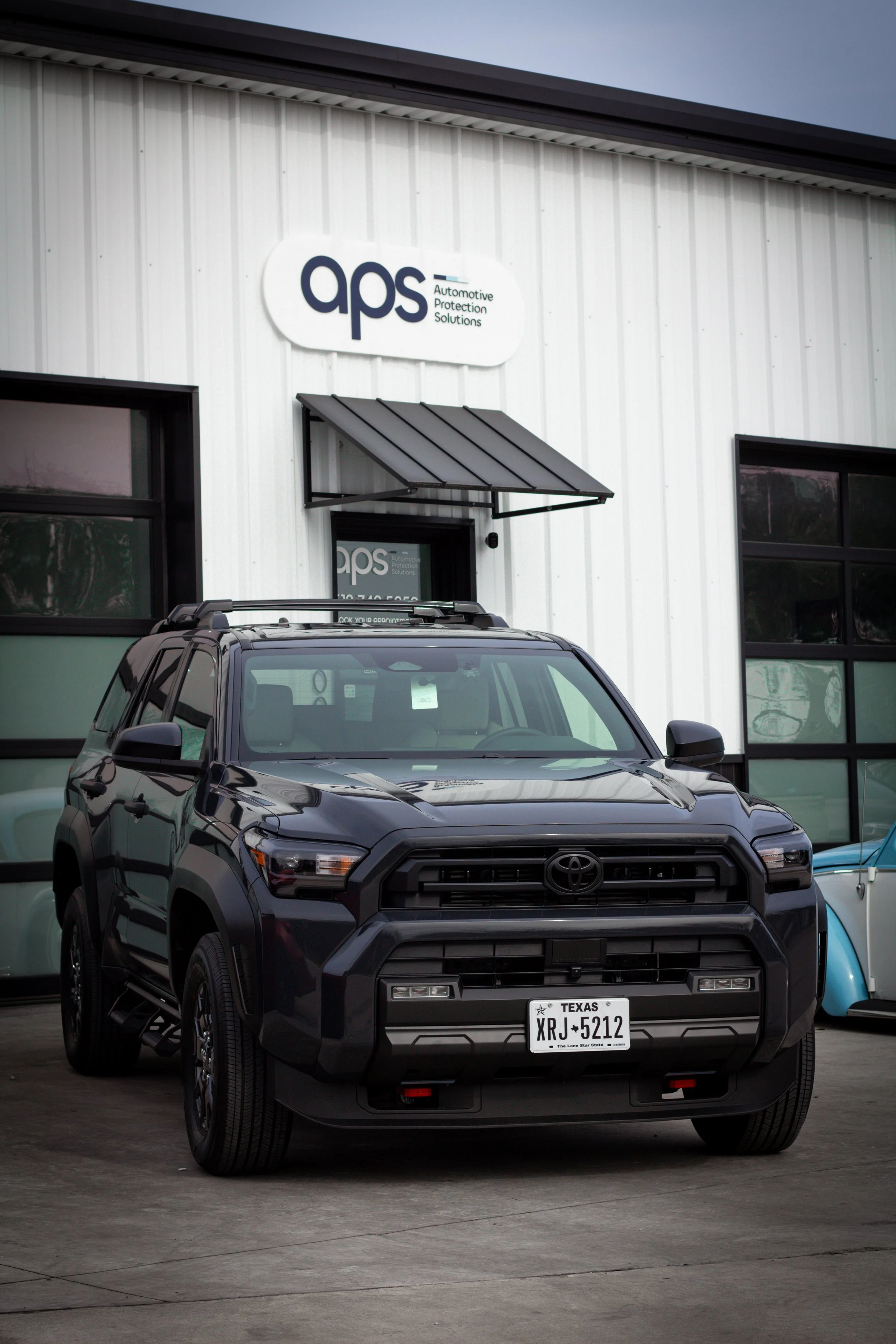 Un automóvil Toyota negro estacionado frente a un edificio con letrero de 'aps' y palabras 'Automotive Protection Solutions' en letras pequeñas.