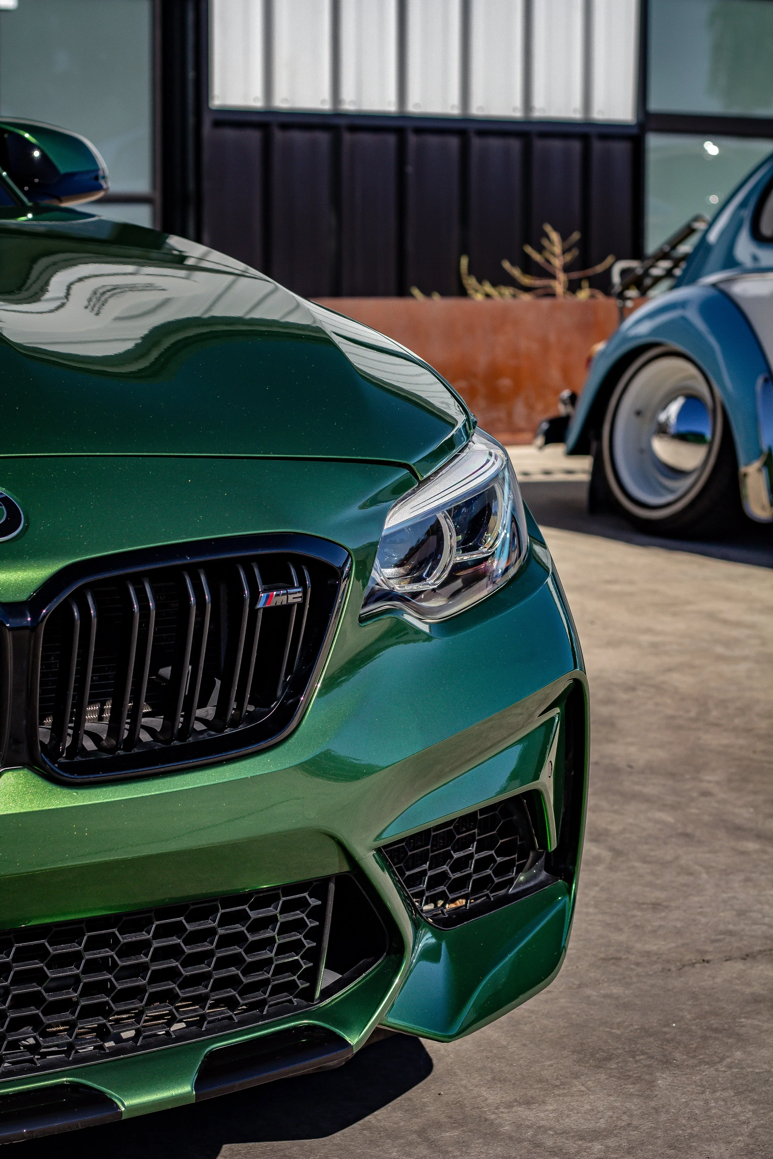 Primer plano de un coche deportivo verde con detalles en negro, en un estacionamiento, con otro coche clásico azul visible al fondo.